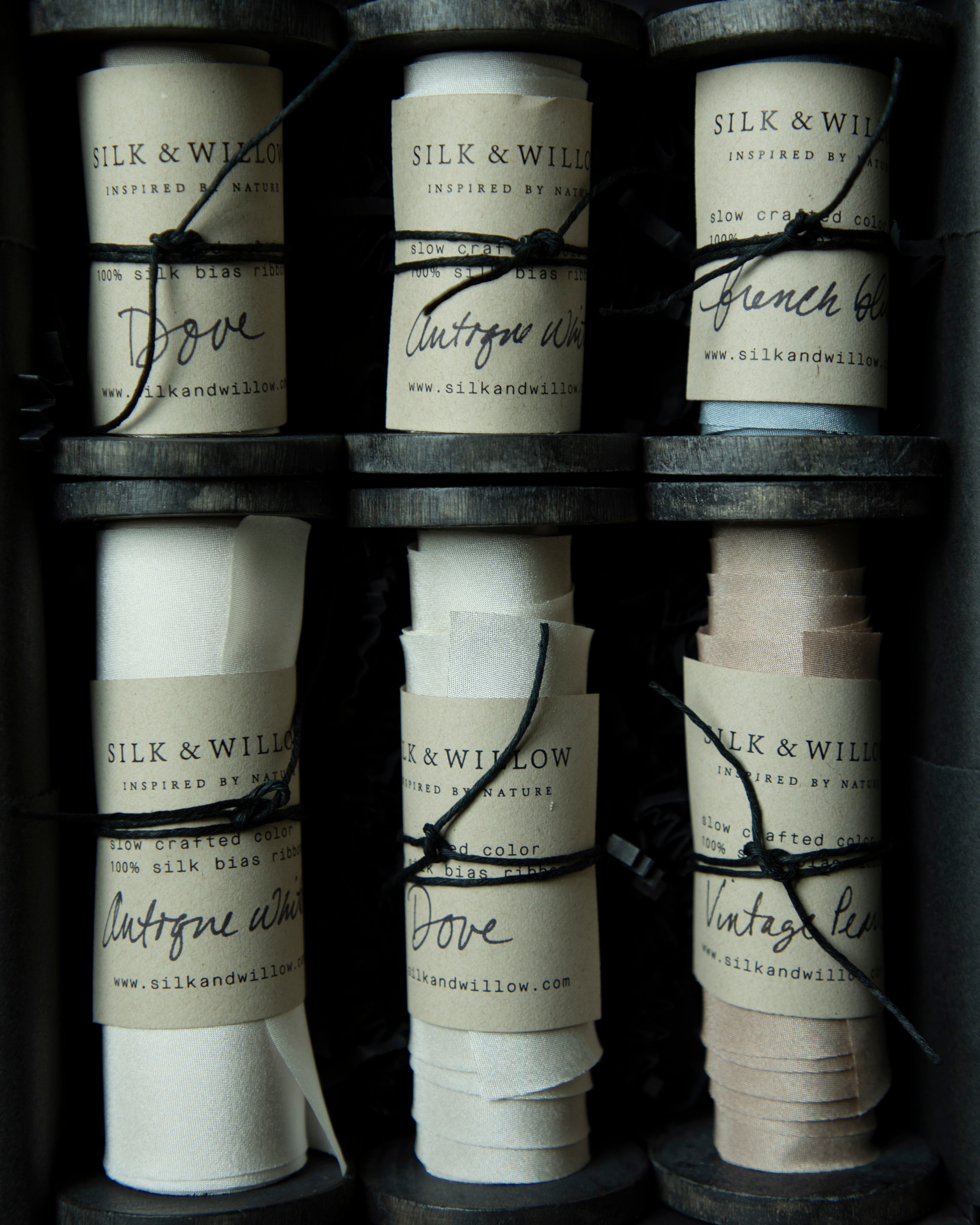 Six rolls of silk ribbon in different colors and widths arranged in a dark gift box. Each roll has a paper label with the brand name 'SILK & WILLOW' and a black string tied around it.