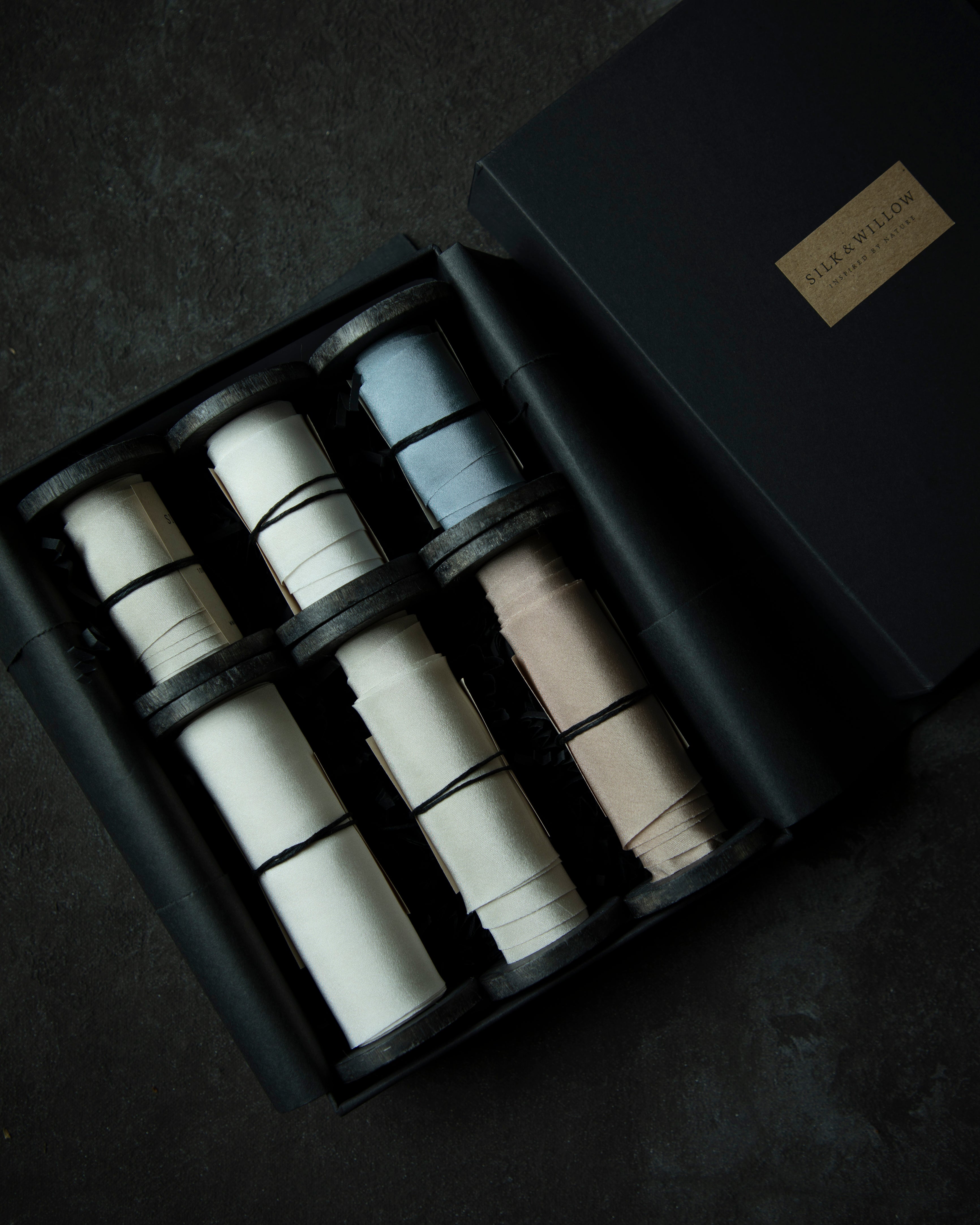 A luxury gift set of six premium ribbons in a black presentation box, shown from a top-down angle on a dark textured surface. The ribbons come in white, light blue, and light beige colors, with a gold-toned brand plaque on the lid.