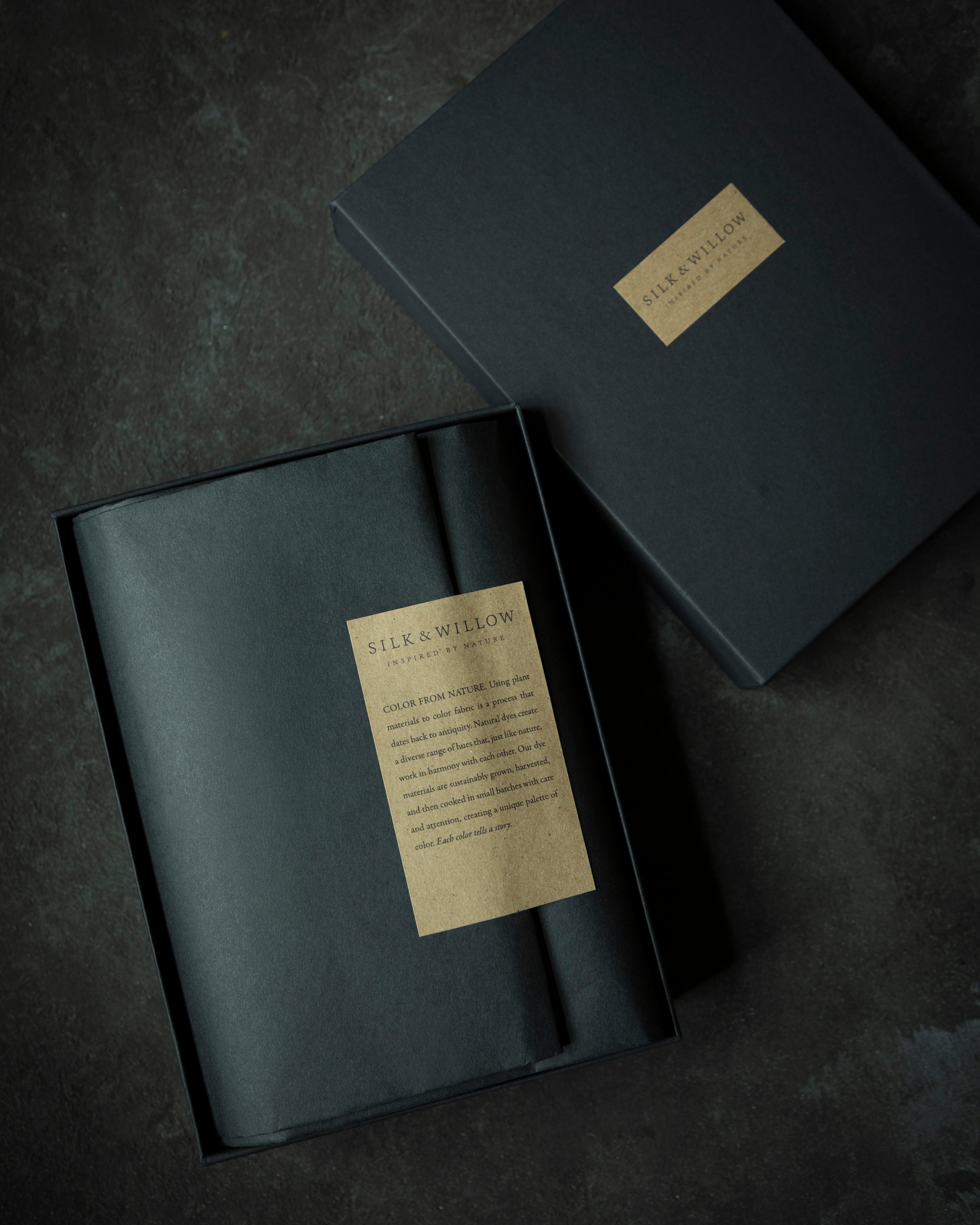 A dark charcoal gray presentation box with a gold label reading 'SILK & WILLOW' and 'The Art of Packaging', shown open with its lid placed beside it on a dark textured background.
