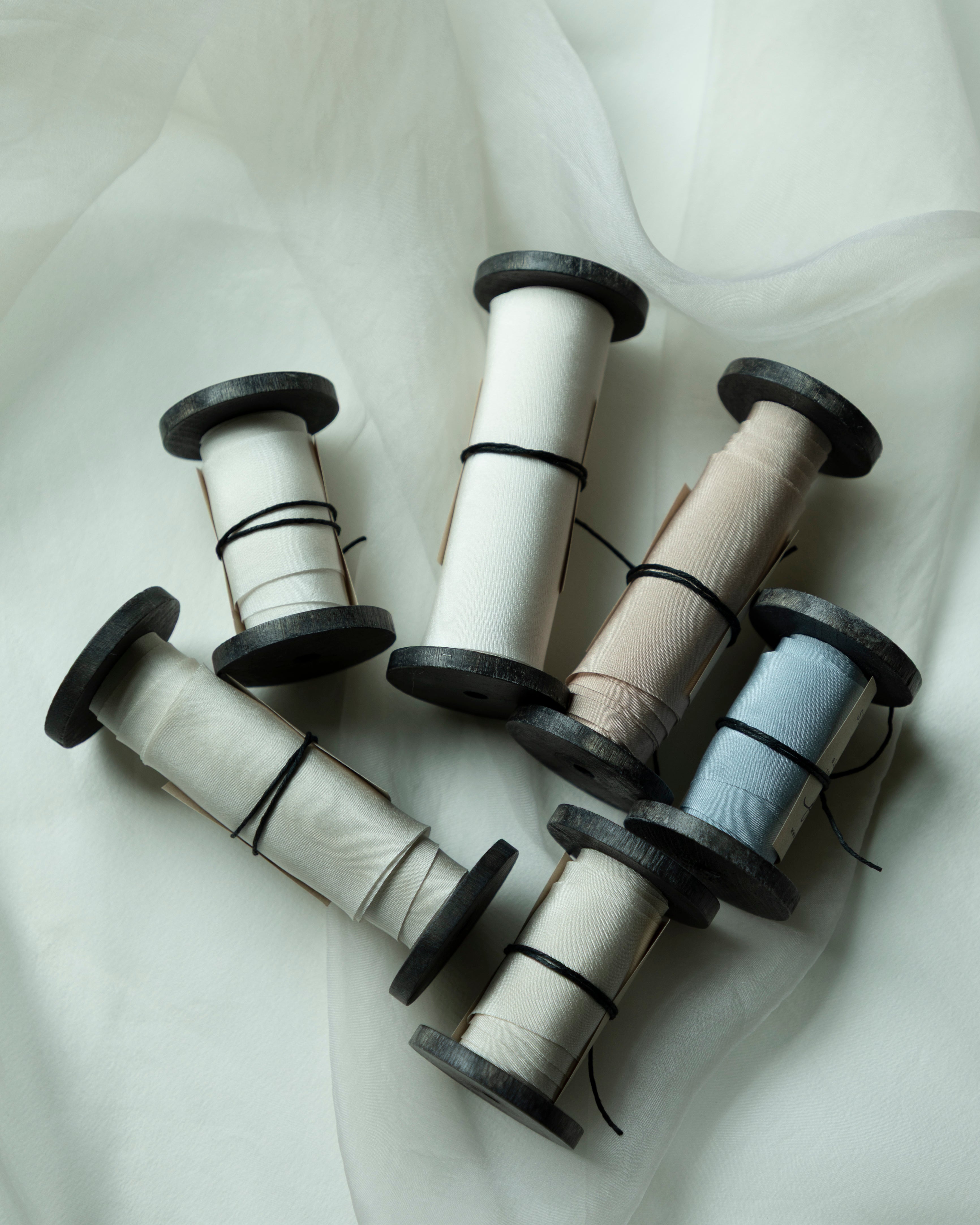Five spools of bridal bouquet silk ribbon in neutral and pastel shades, including cream, beige, light gray, and pale blue, arranged on a draped white fabric background.