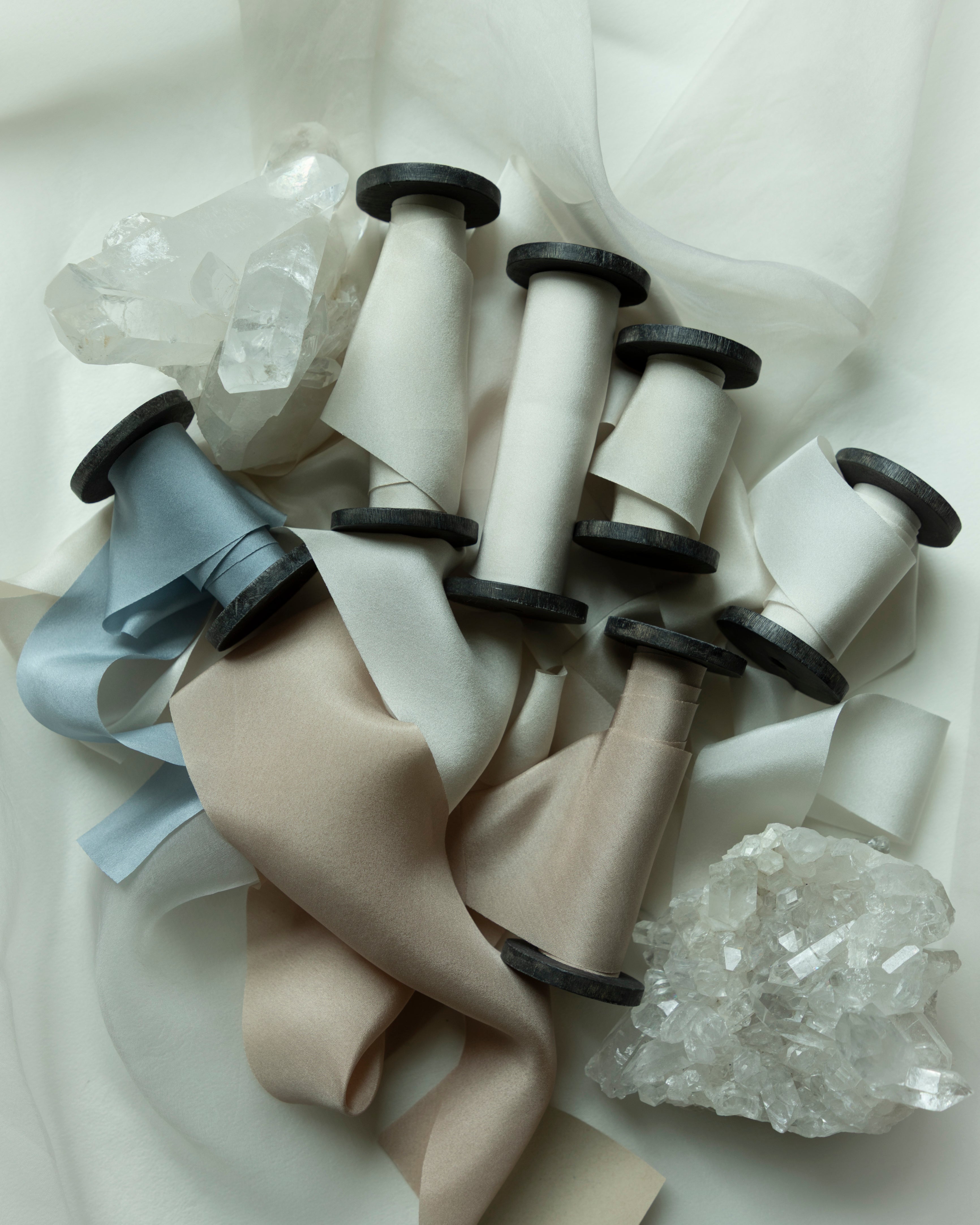 A styled flat-lay of multiple spools of hand-dyed silk ribbon in neutral tones, including beige, cream, light blue, and taupe, arranged on a draped white fabric background with clear crystal clusters.