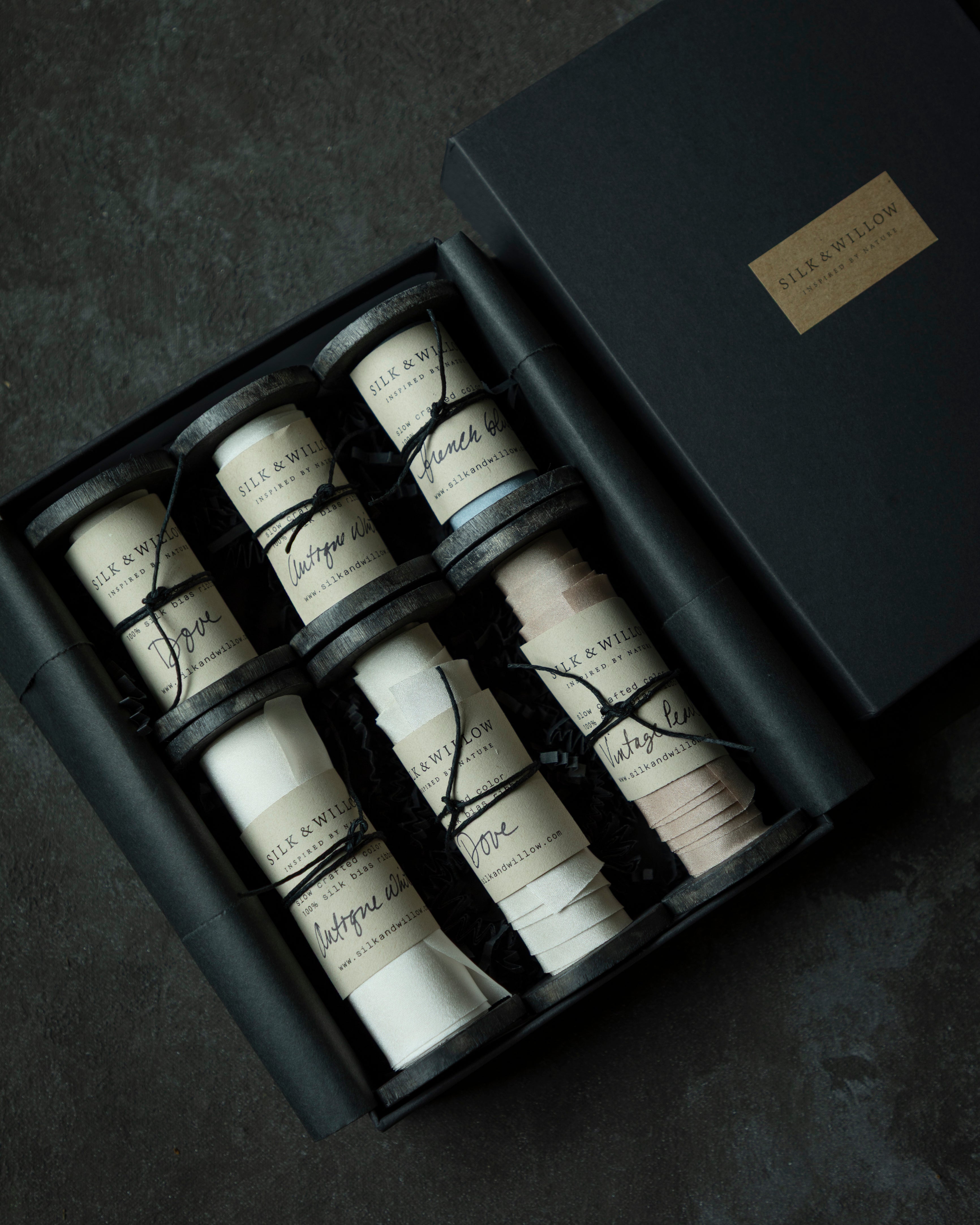 An open black gift box containing six spools of premium silk ribbon, shown on a dark textured surface. The closed matching box with a gold label sits beside it.