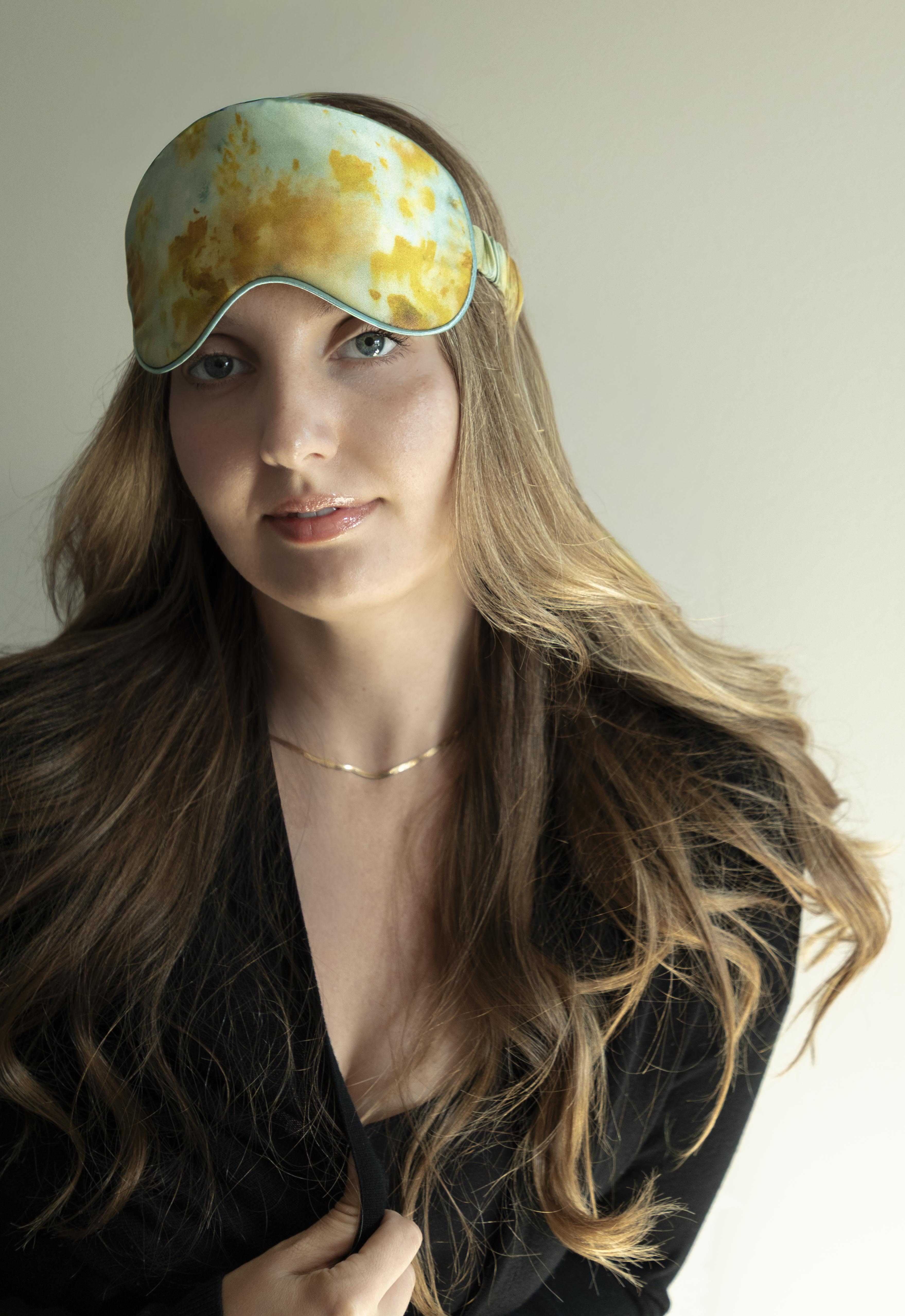 Woman wearing a yellow and green patterned sleep mask against a neutral background