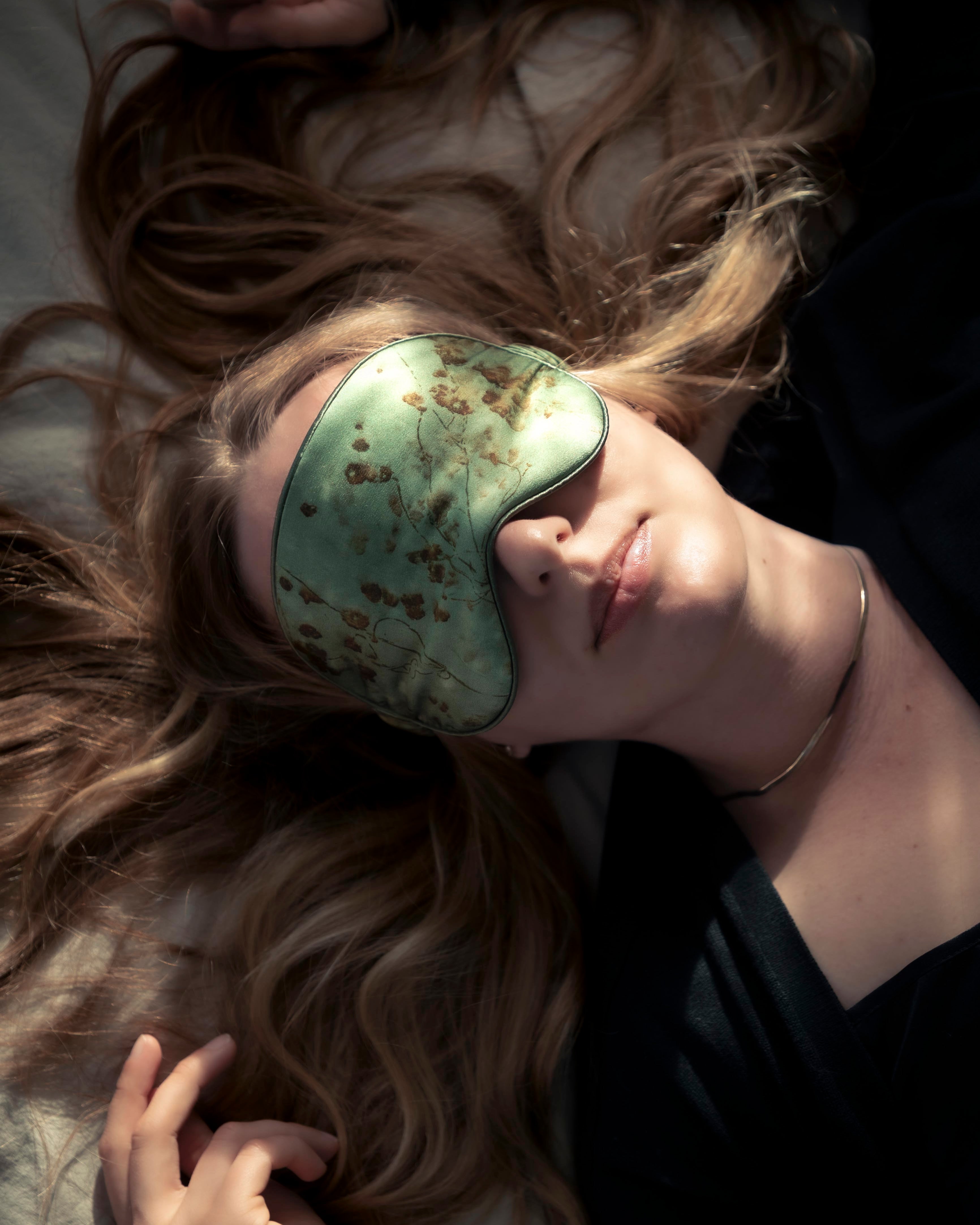a woman lying on a bed in the sunlight wearing a botanical print organic silk sleep eye mask