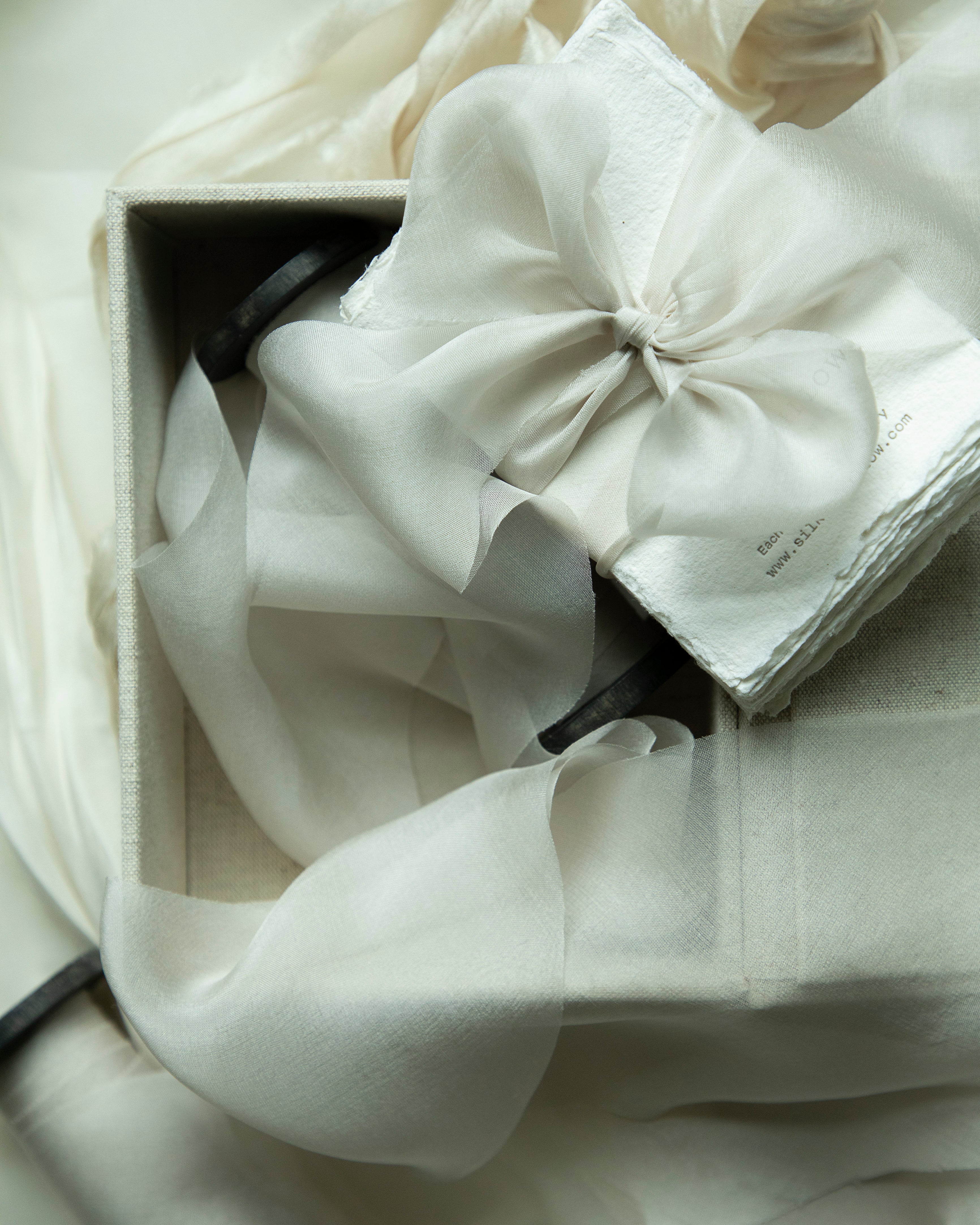 White fabric with a large bow in a box on a light background. A small stack of textured paper wrapped in a soft, pale ribbon, resting on a light fabric backdrop.
