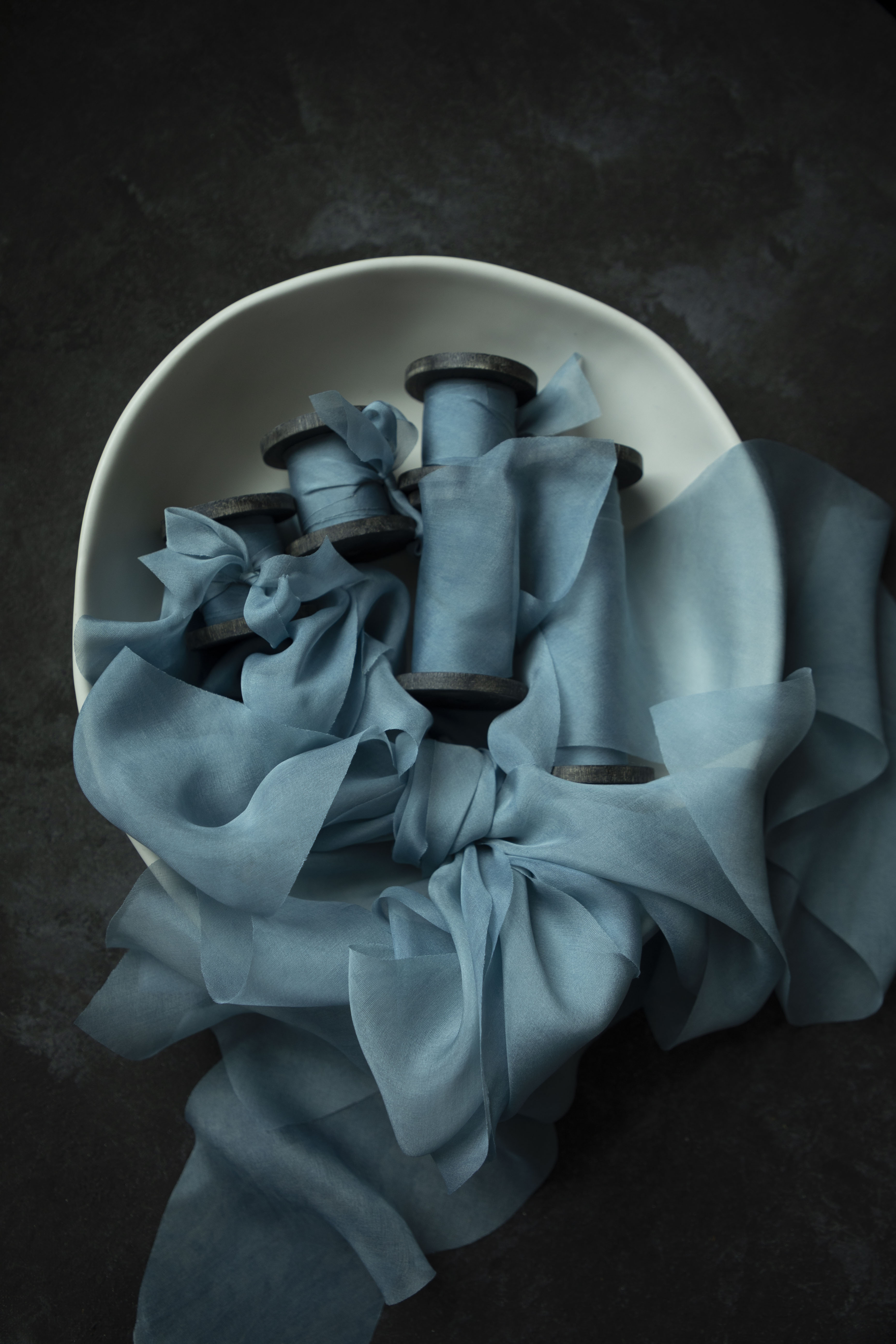 Blue ribbon, indigo dyed, wrapped on antique wood spools in a white bowl on a dark background