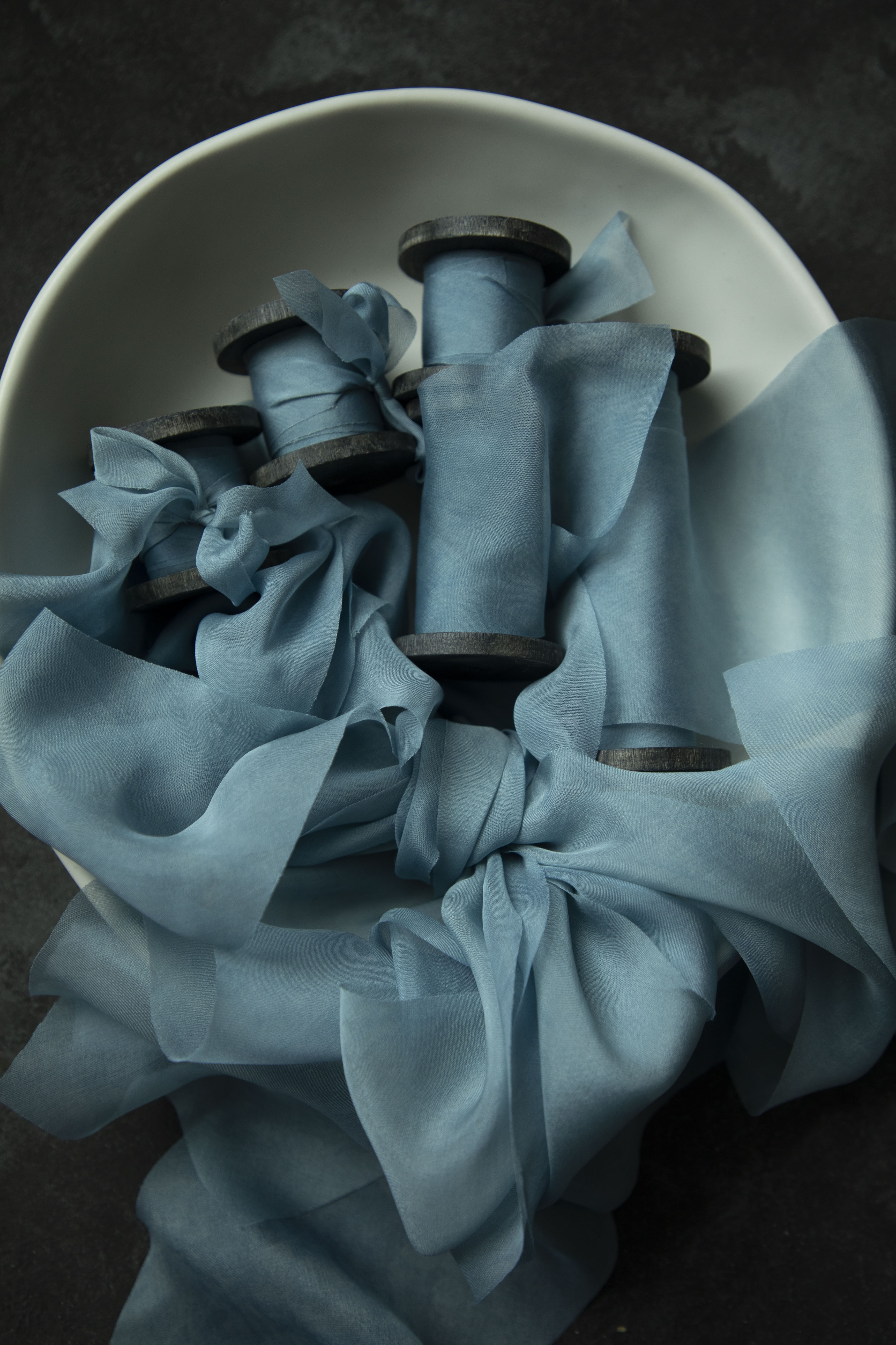 Duchess Blue silk ribbon wrapped around wooden spools inside a white bowl on a dark surface