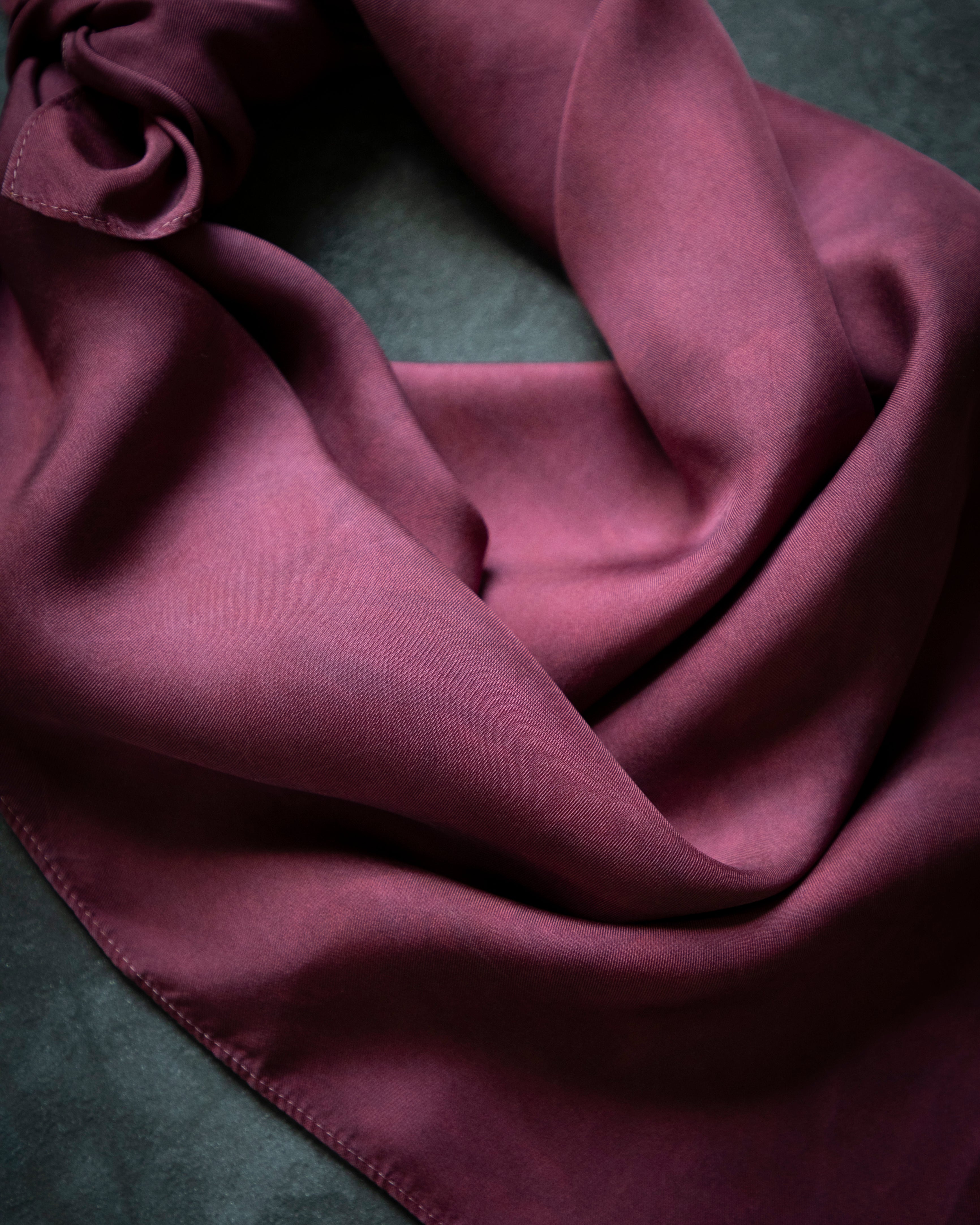 A draped mauve-purple scarf with a satin-like sheen, styled in soft folds on a dark textured background.