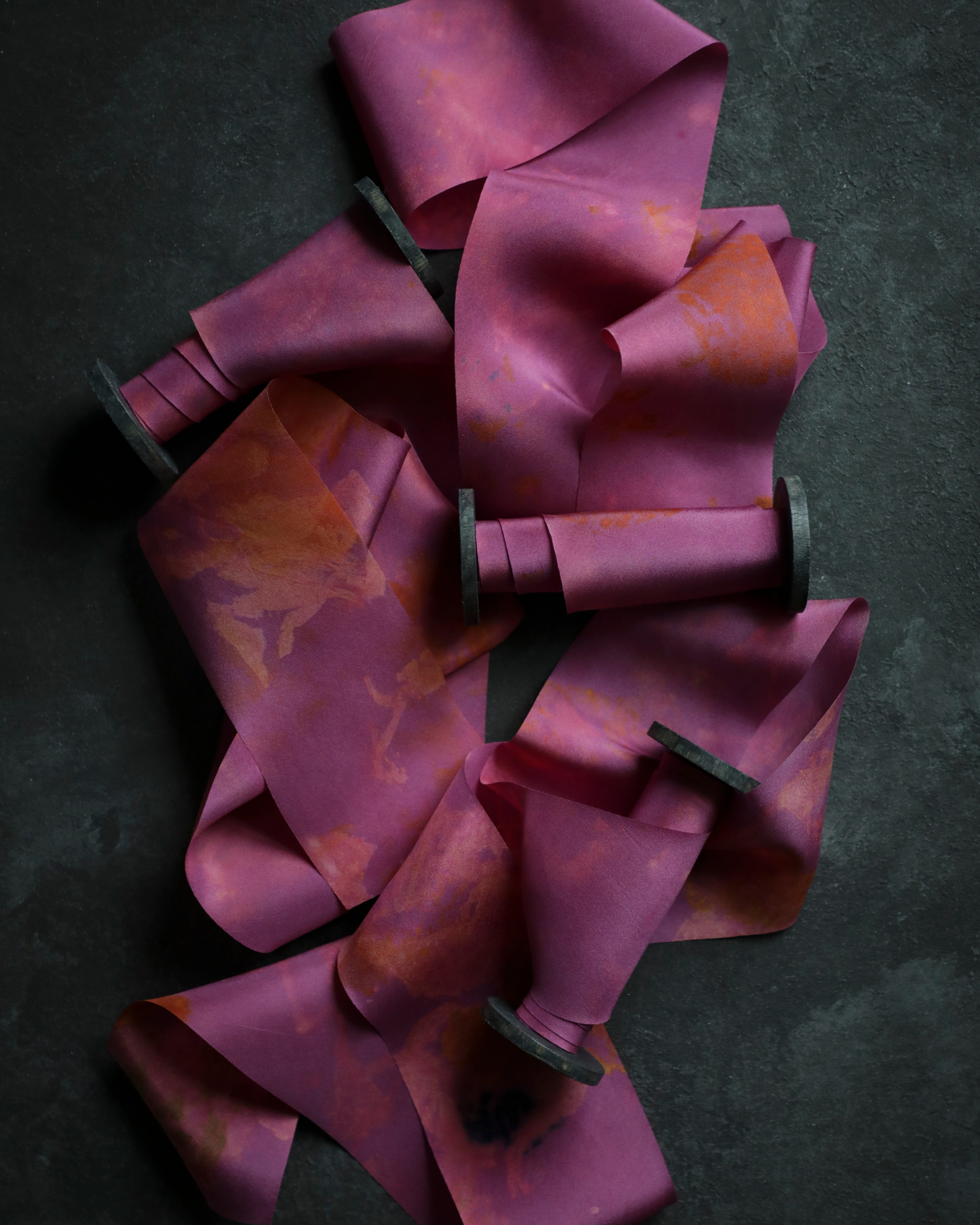 A collection of silk ribbons with eco-printed botanical patterns in fuchsia, magenta, and other colors, laid out on a dark textured background.