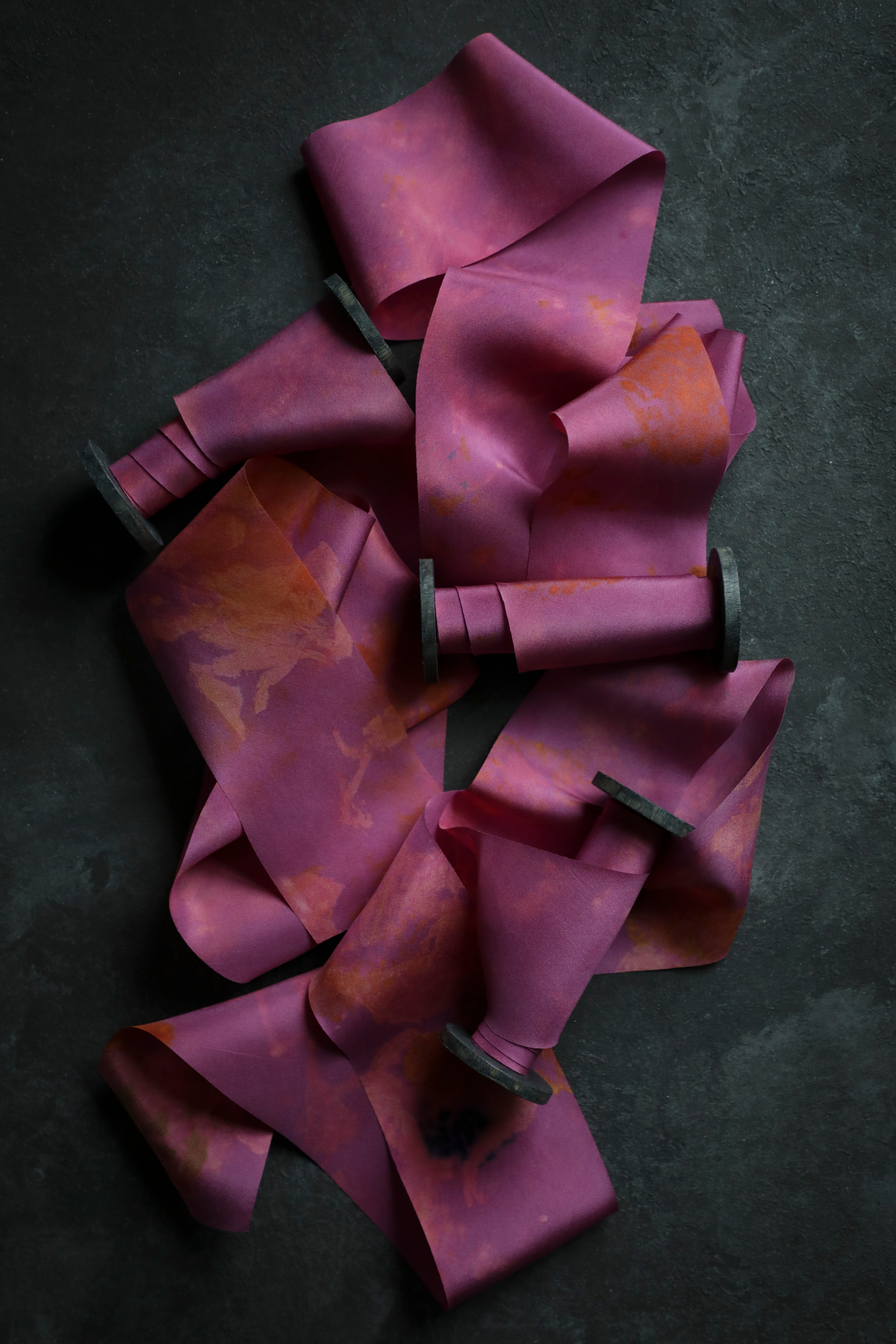 A collection of silk ribbons with eco-printed botanical patterns in fuchsia, magenta, and other colors, laid out on a dark textured background.