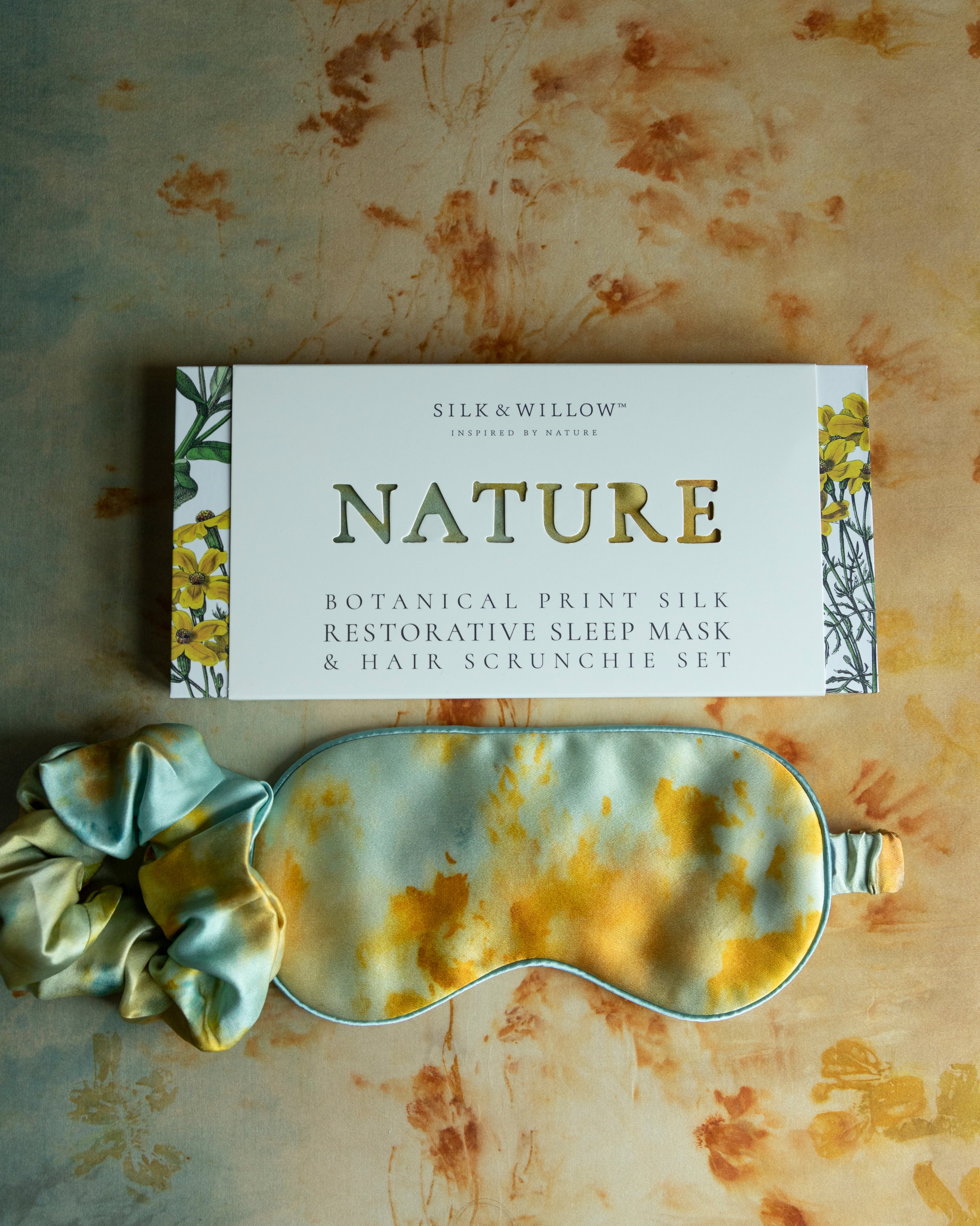 A botanical print silk sleep eye mask and hair scrunchie set in teal and marigold colors, presented on a cardboard packaging with 'NATURE' text.