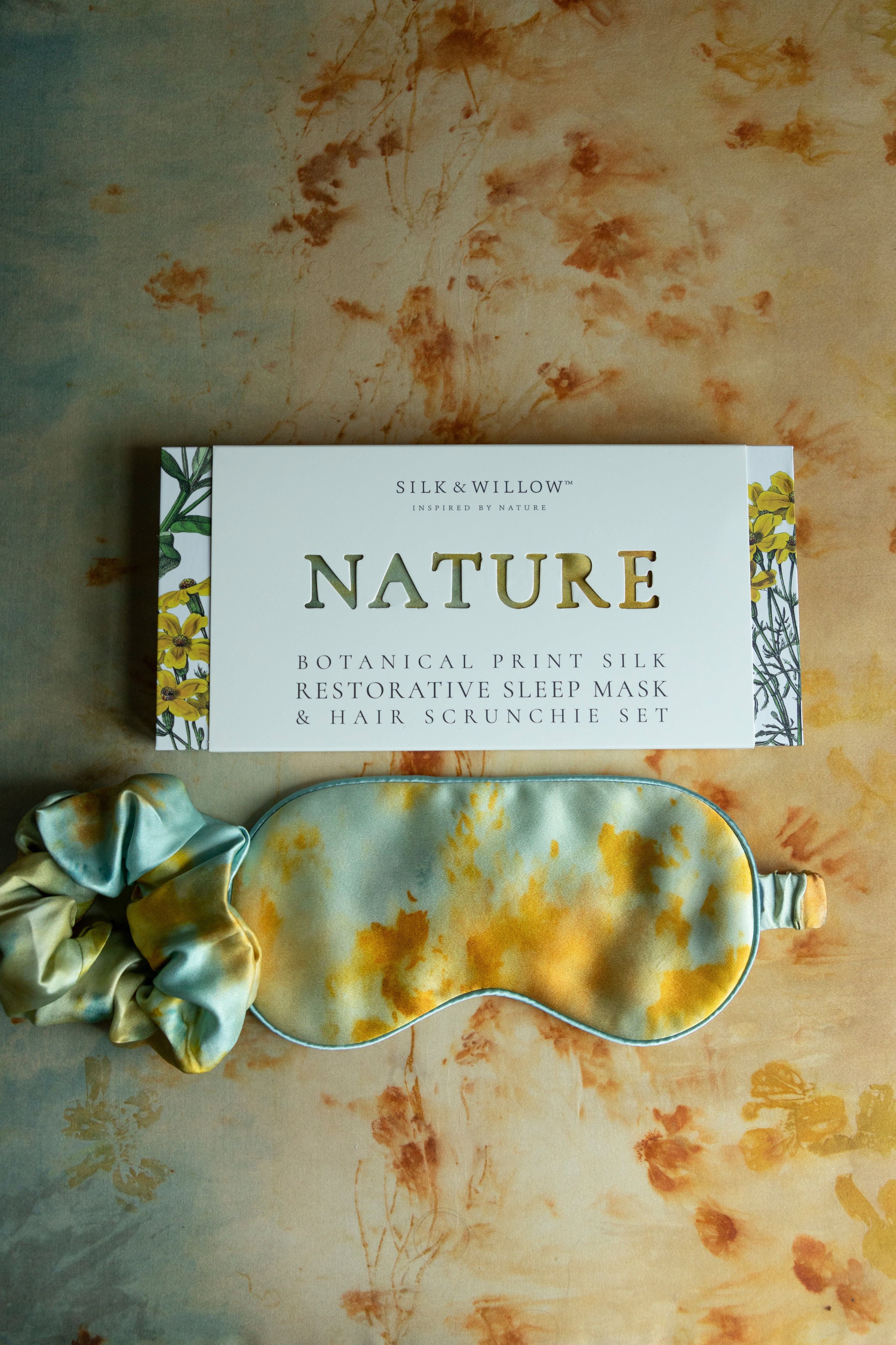 A botanical print silk sleep eye mask and hair scrunchie set in teal and marigold colors, presented on a cardboard packaging with 'NATURE' text.