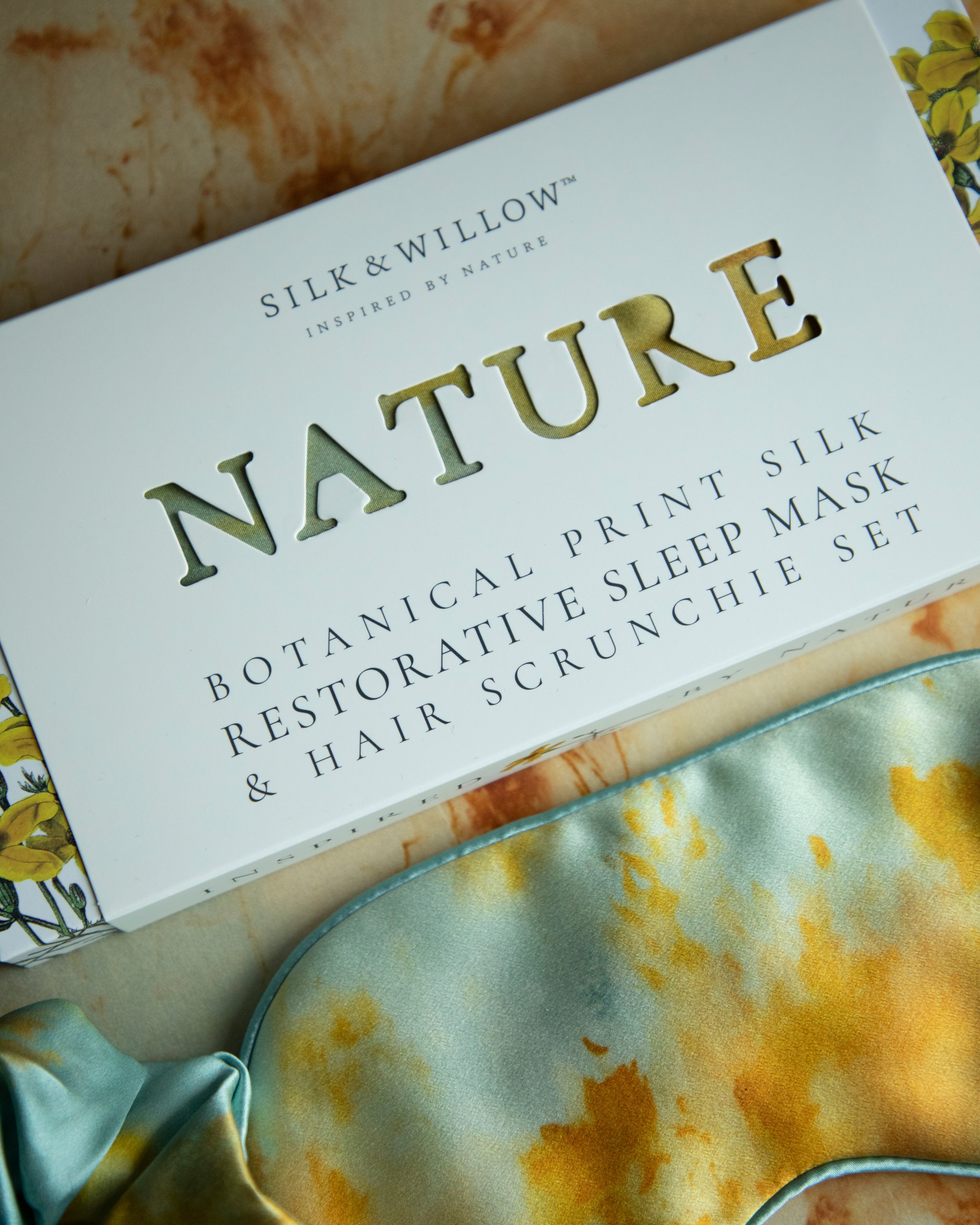 Silk & Willow Nature botanical print silk restorative sleep mask and hair scrunchie set on a textured surface.