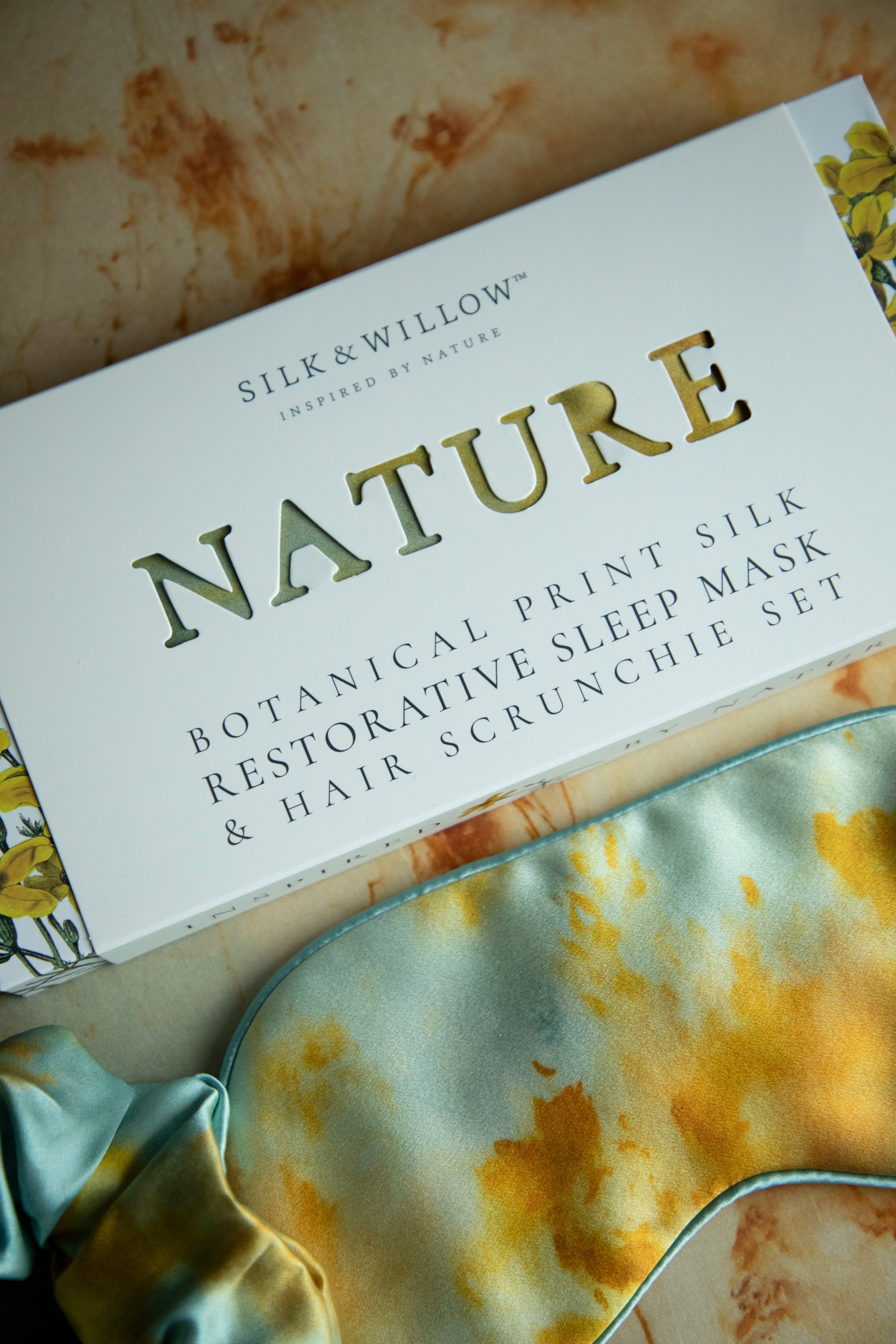Silk & Willow Nature botanical print silk restorative sleep mask and hair scrunchie set on a textured surface.