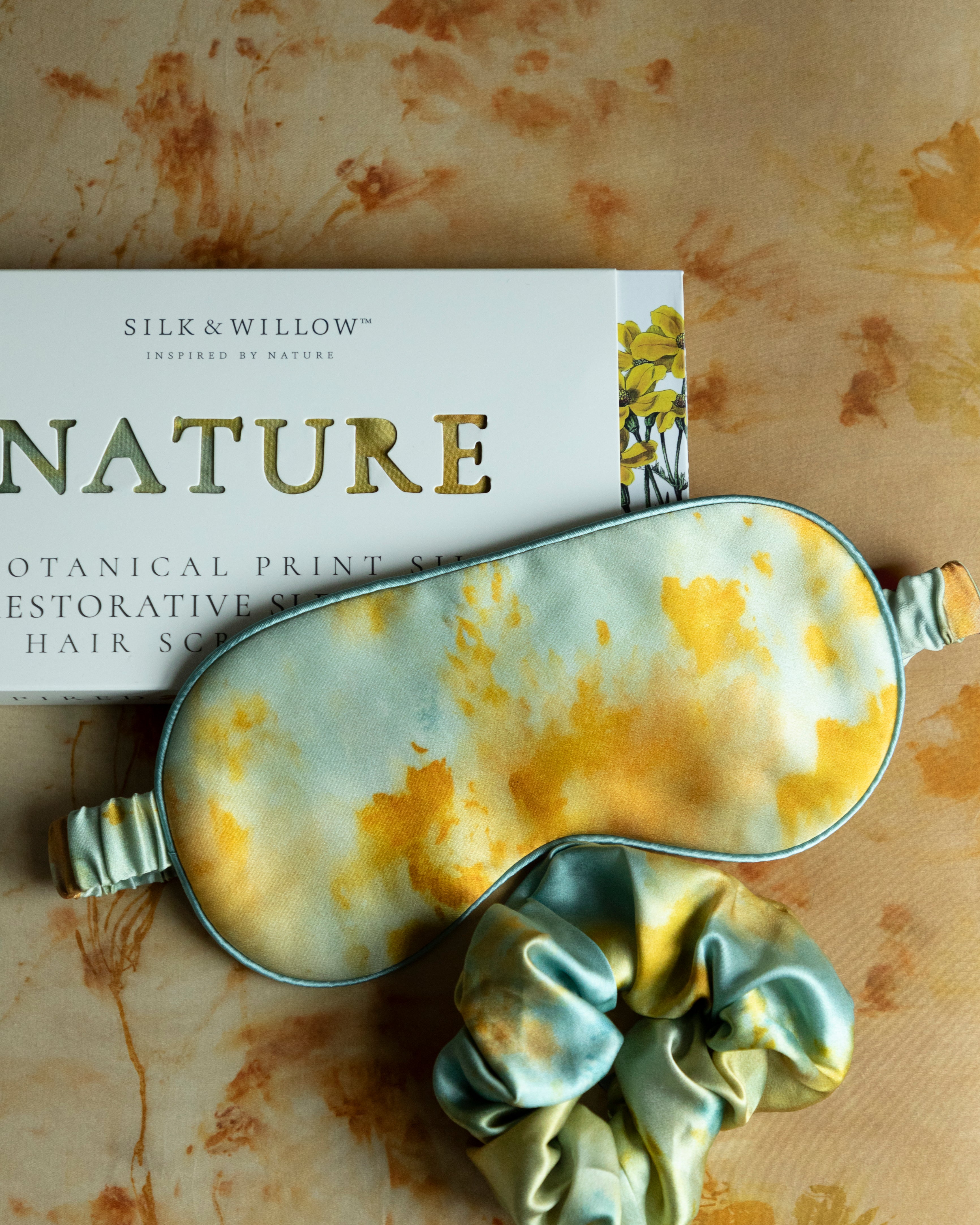 Silk & Willow Nature botanical print silk restorative sleep mask and hair scrunchie set on a textured surface.