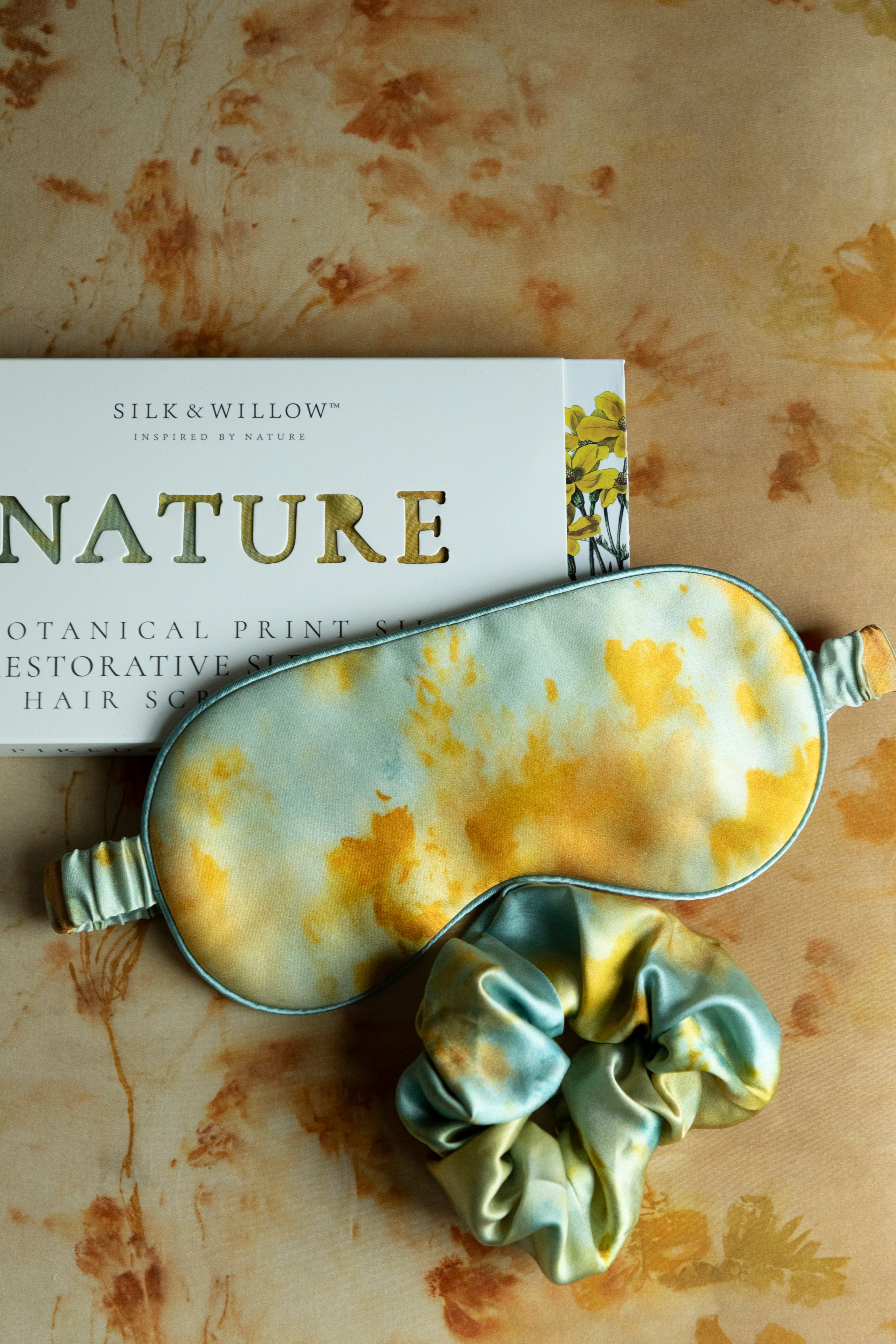 Silk & Willow Nature botanical print silk restorative sleep mask and hair scrunchie set on a textured surface.