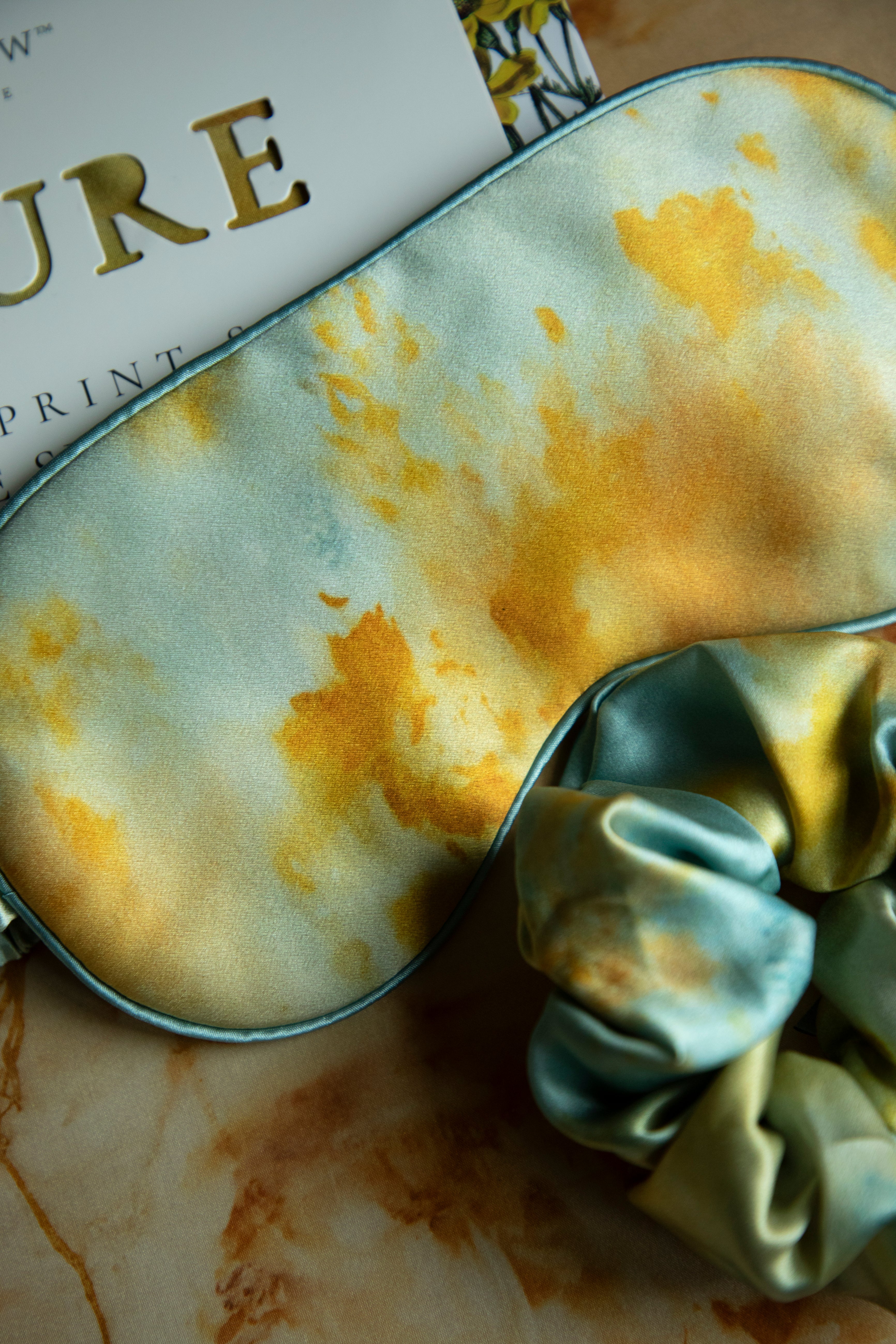 Silk & Willow Nature botanical print silk restorative sleep mask and hair scrunchie set on a textured surface.