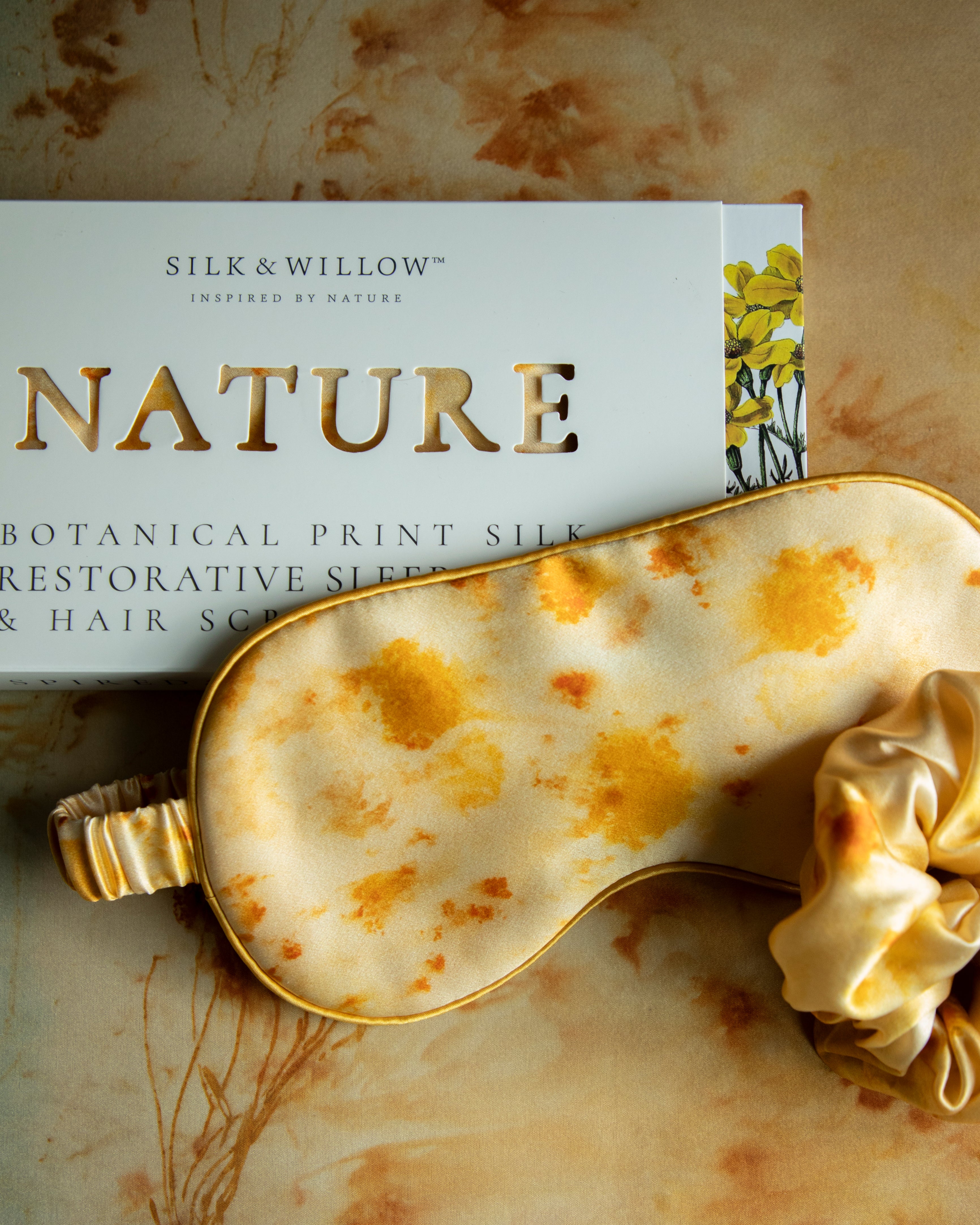 A silk sleep eye mask with a botanical print in marigold yellow, displayed next to its packaging, indicating a luxurious and eco-friendly product.
