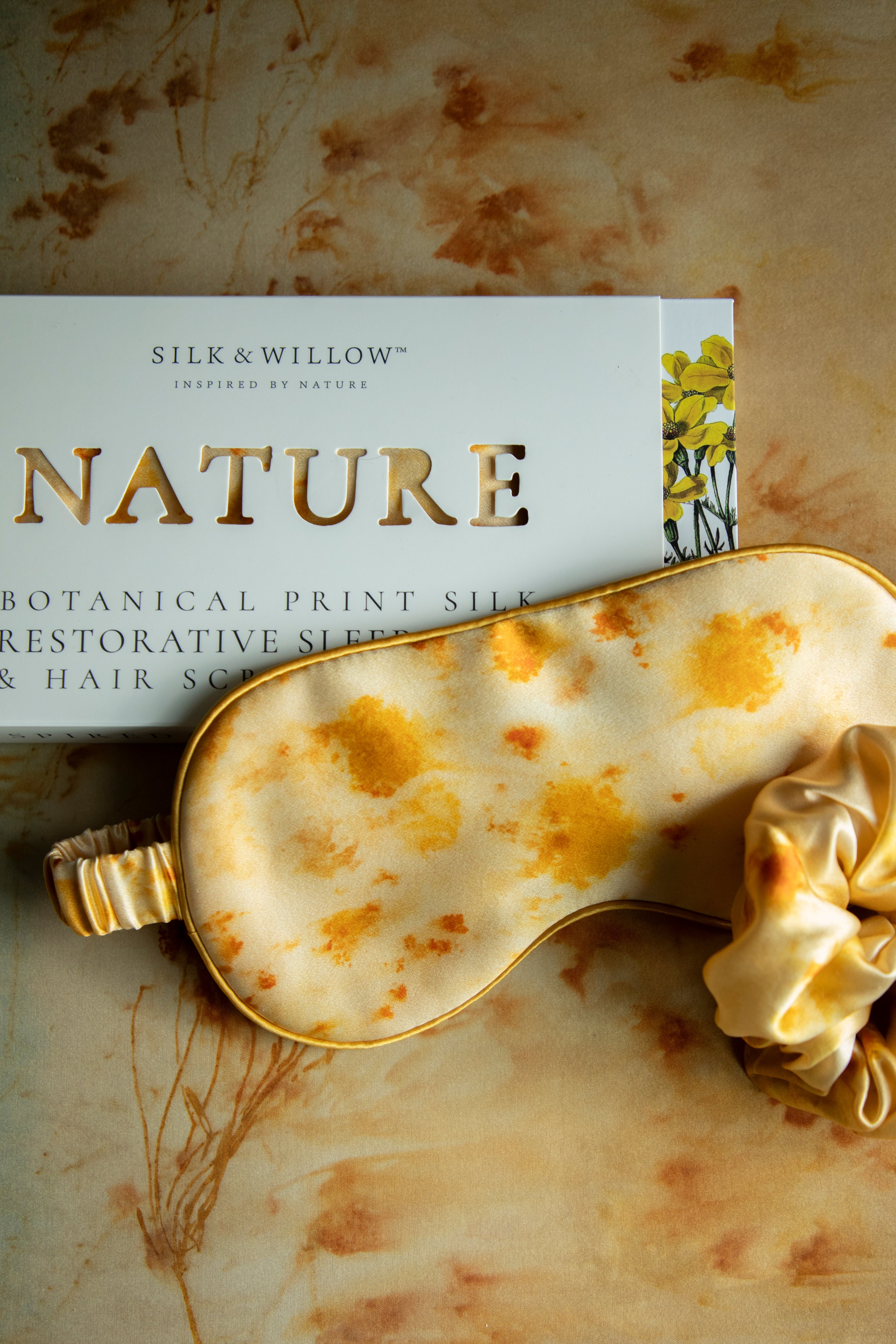 A silk sleep eye mask with a botanical print in marigold yellow, displayed next to its packaging, indicating a luxurious and eco-friendly product.