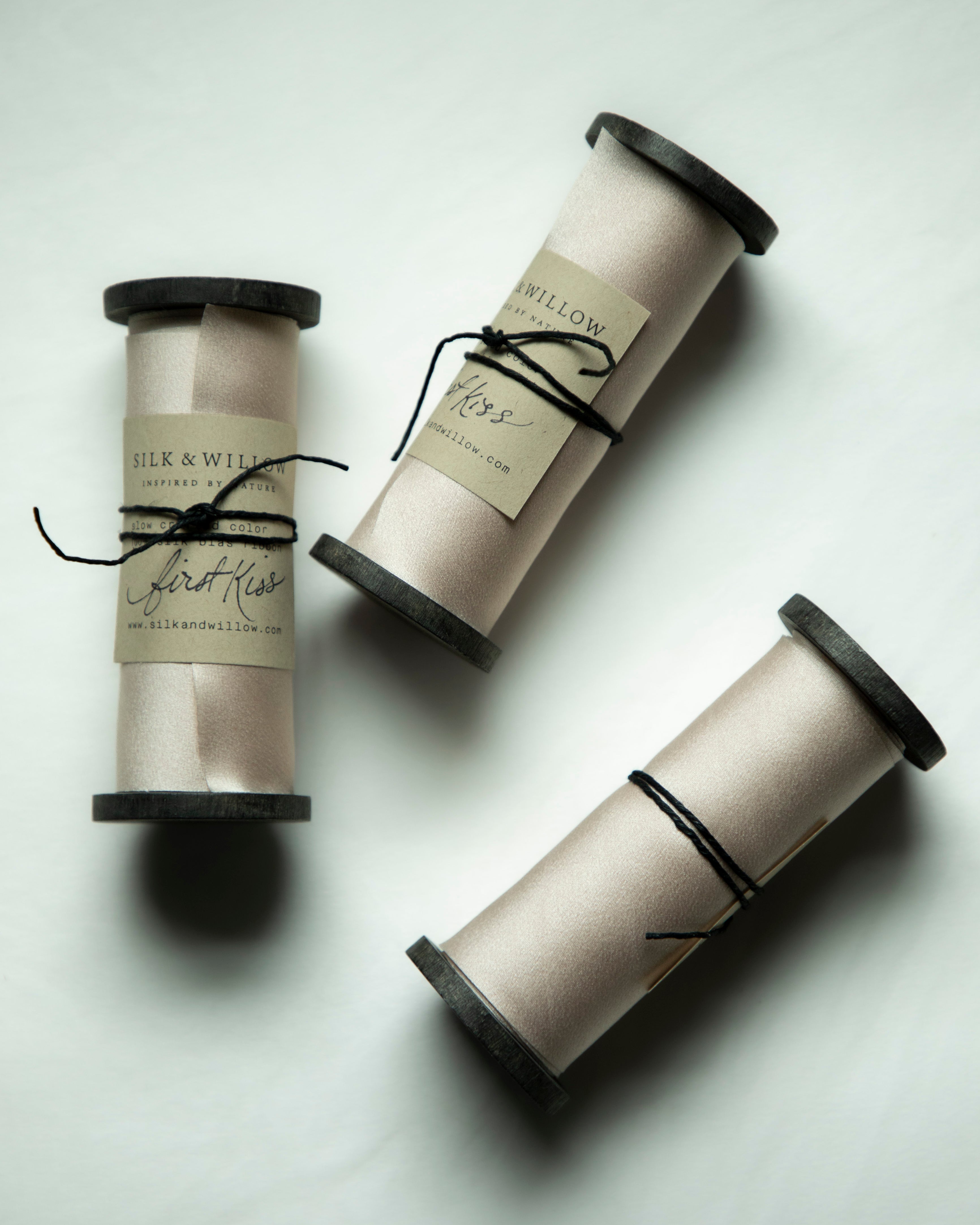 Three spools of silk ribbon in a light blush color on a black wood spool, tied with black string and featuring silk & Willow labels, arranged on a light gray background.