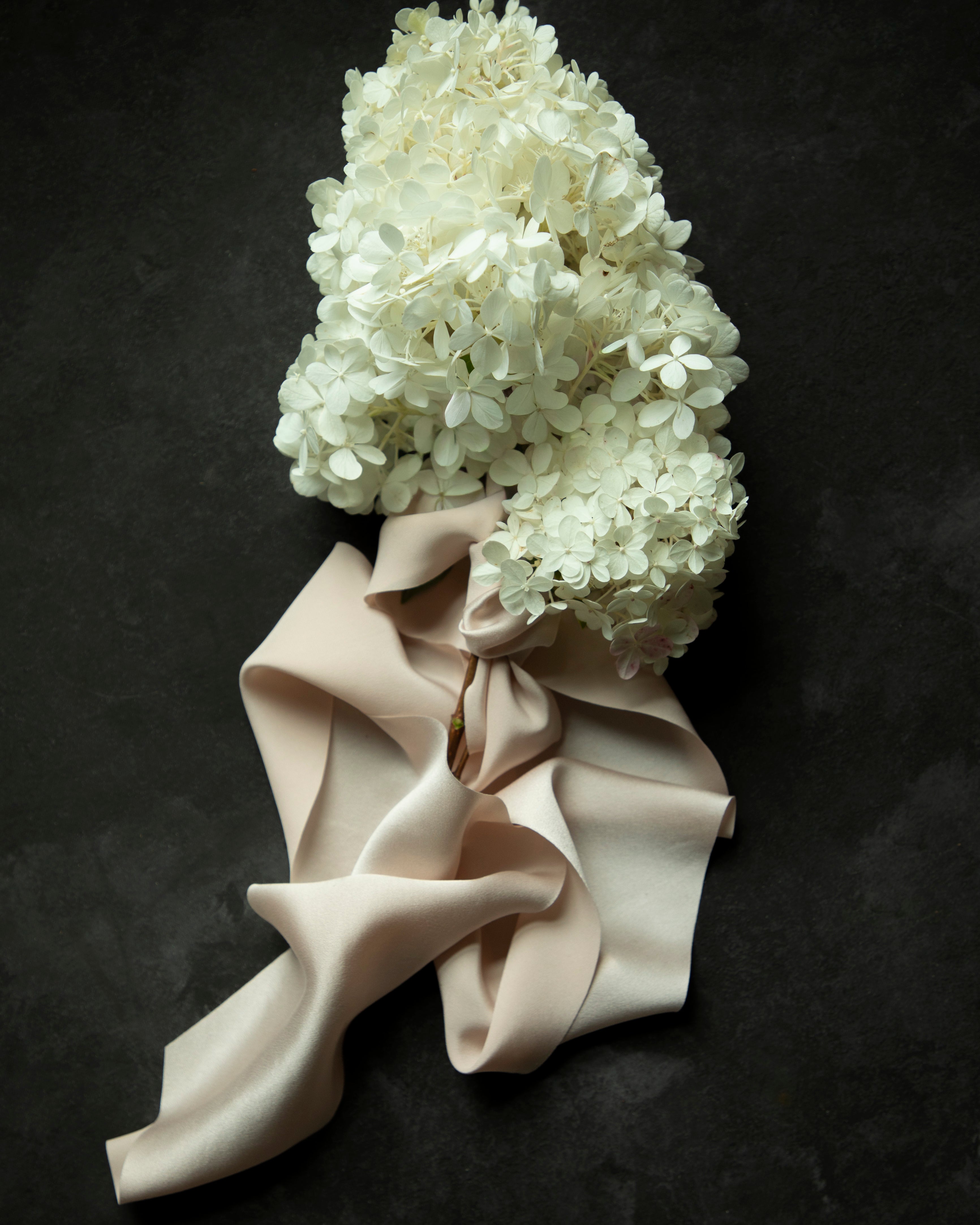 White hydrangea flower bouquet wrapped with a premium bridal silk satin ribbon, laid flat on a dark textured background.