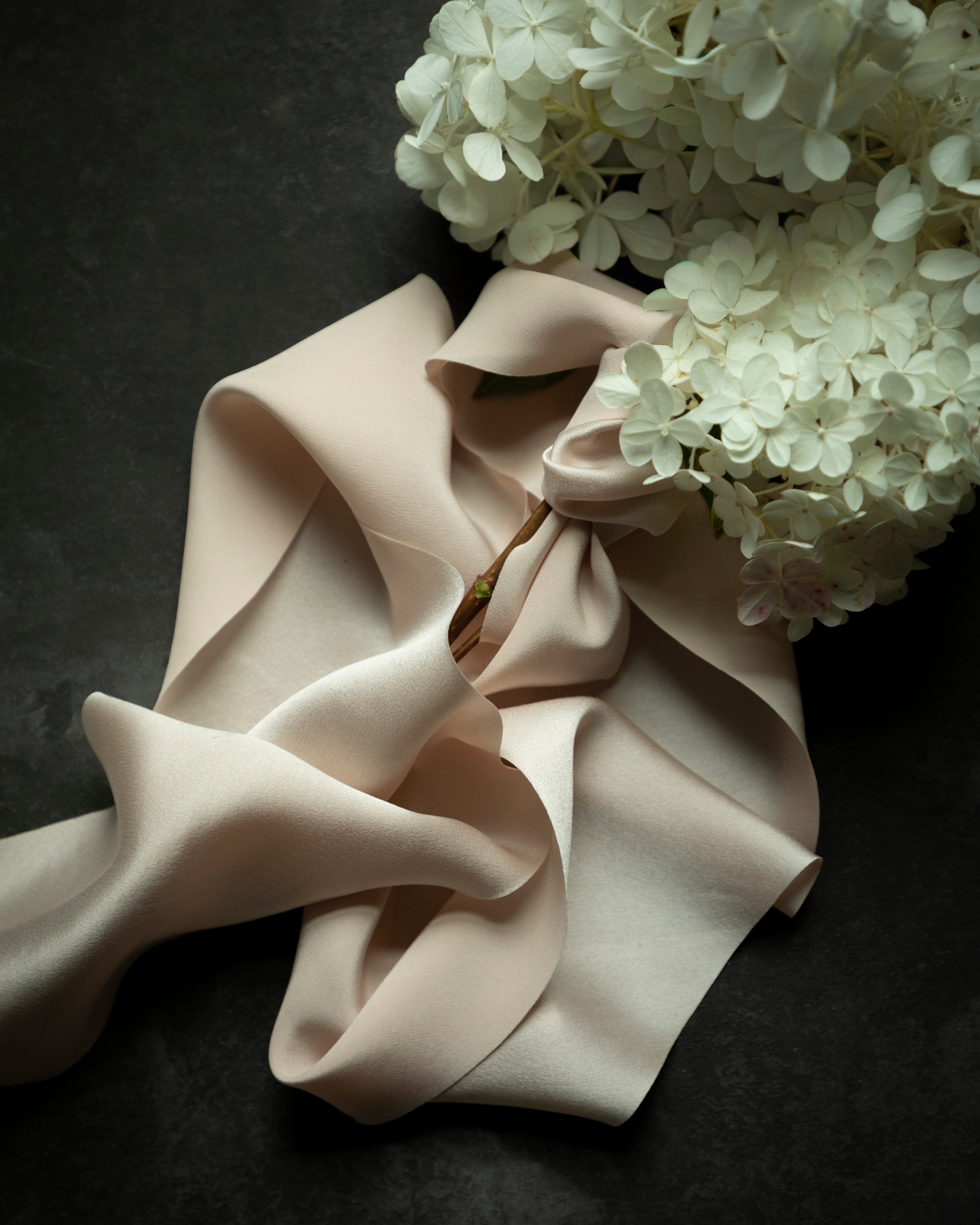 A draped piece of pale blush pink satin ribbon with soft folds, styled with white hydrangea flowers on a dark textured background.