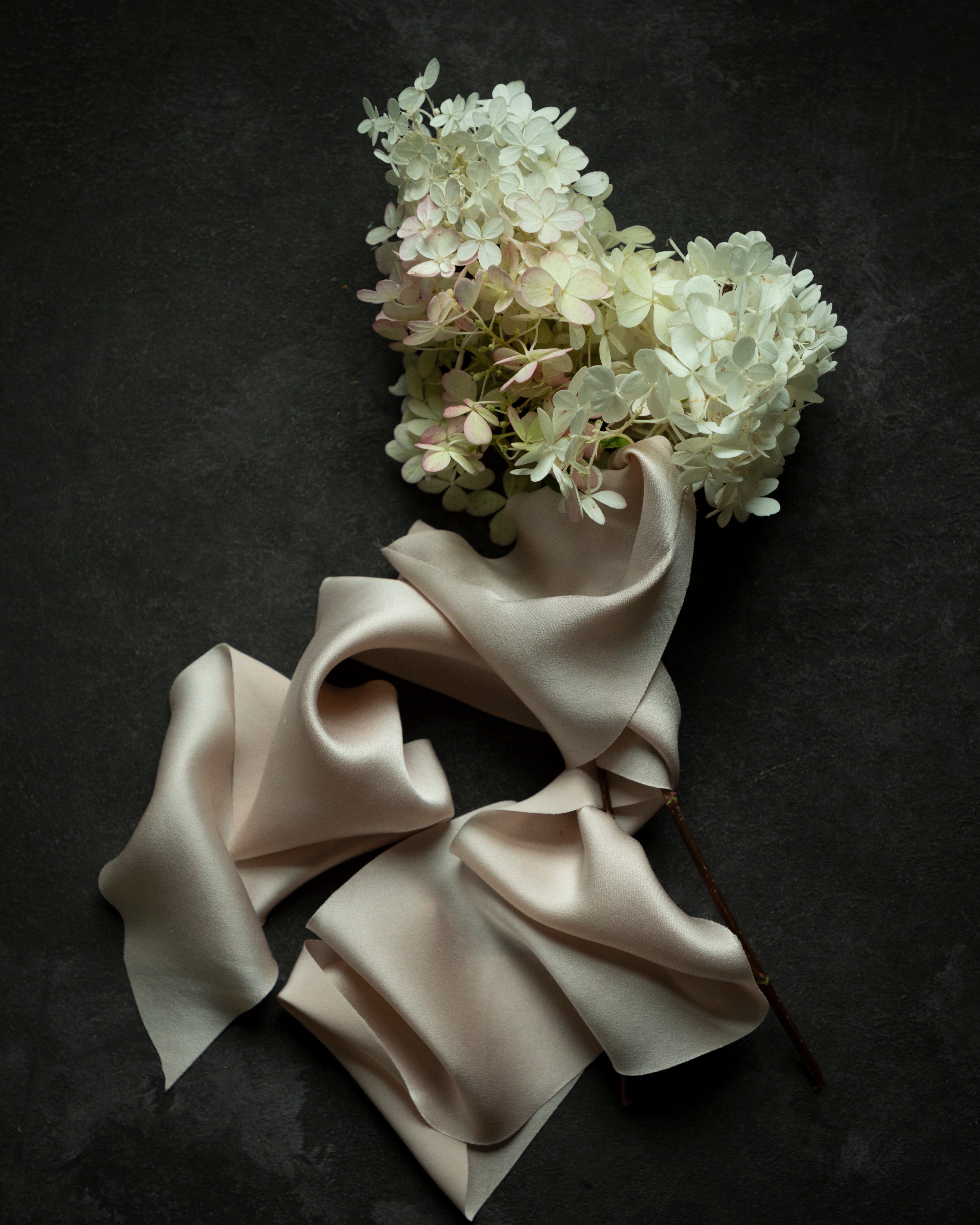 A bouquet of white and pale pink hydrangea flowers with a draped blush silk ribbon, arranged on a dark textured background.
