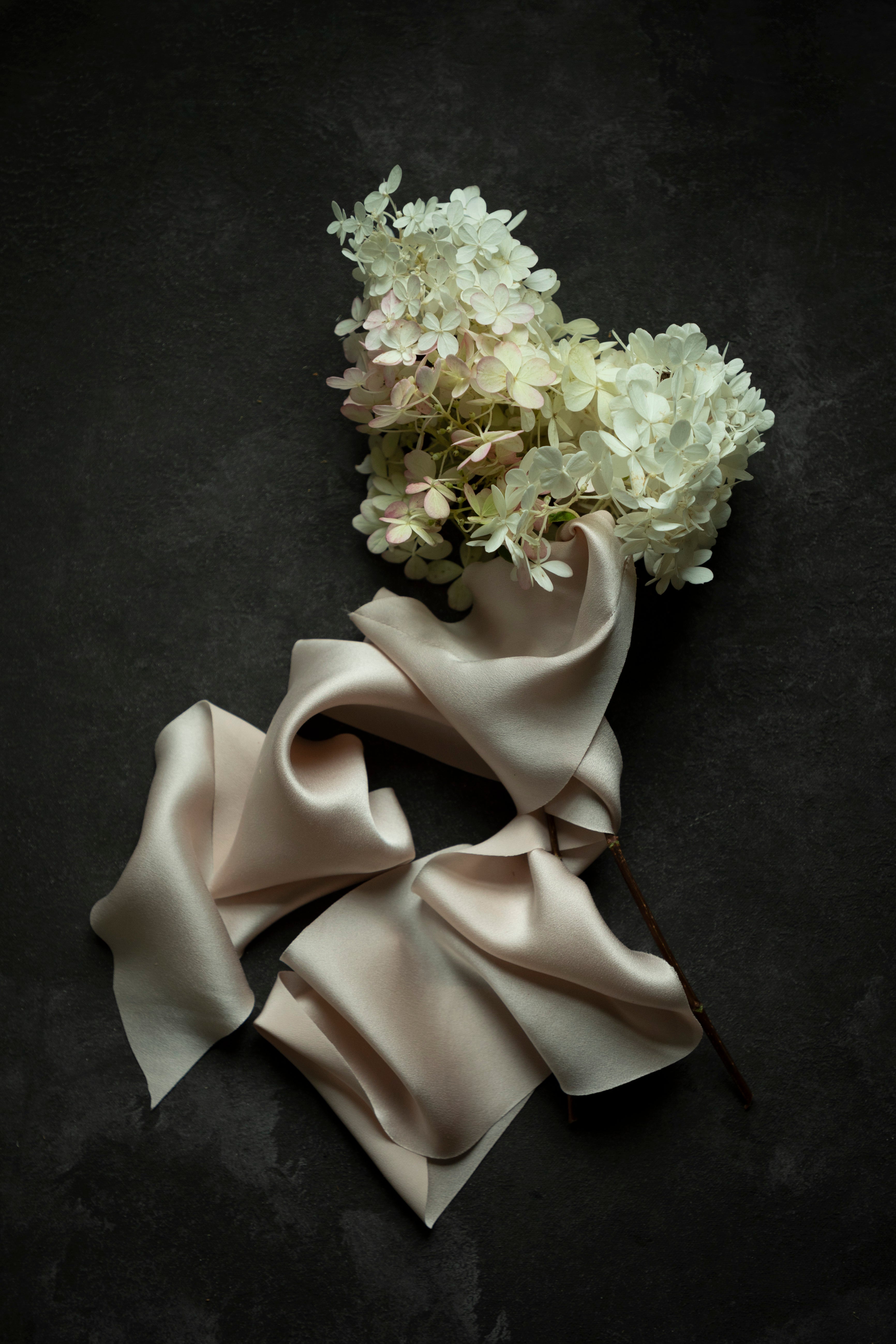 A bouquet of white and pale pink hydrangea flowers with a draped blush silk ribbon, arranged on a dark textured background.