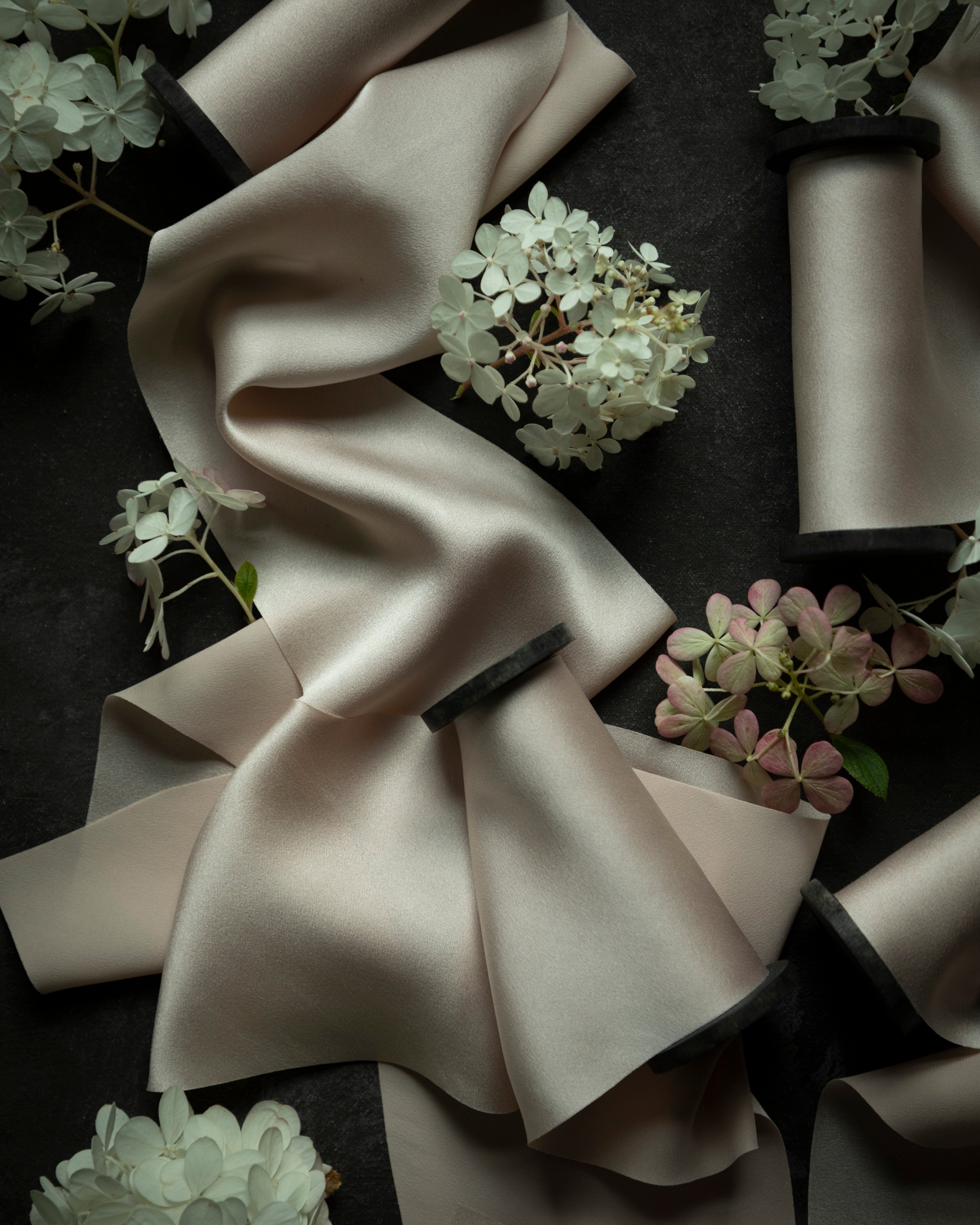 A draped piece of silk satin bridal bouquet ribbon in a light blush color, styled with white and pink hydrangea flowers on a dark textured background.