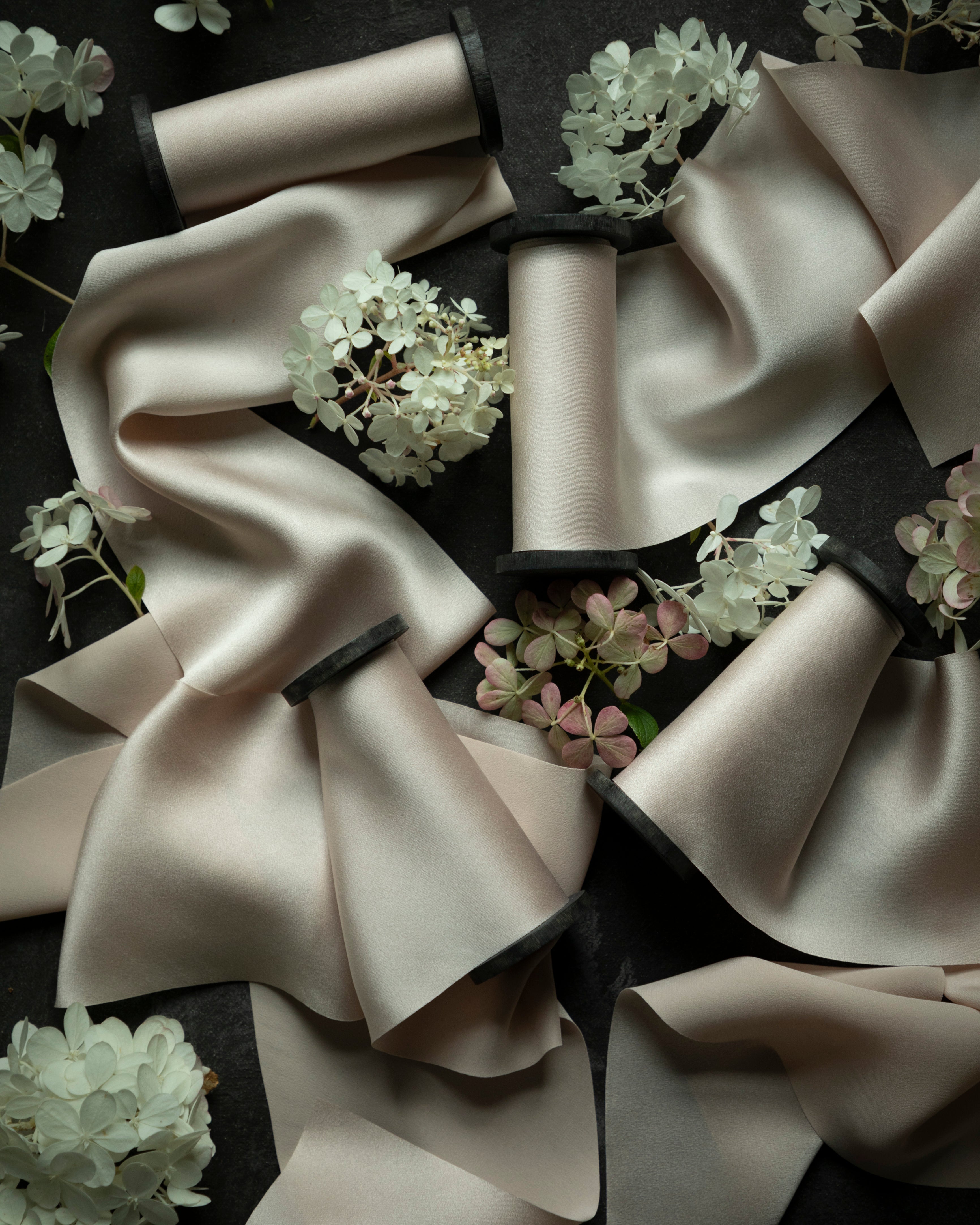 A styled flat-lay of multiple satin ribbon spools arranged on a dark background with decorative white and pale pink flowers. The ribbons are a warm blush-beige tone with dark inner ends visible on the spools.