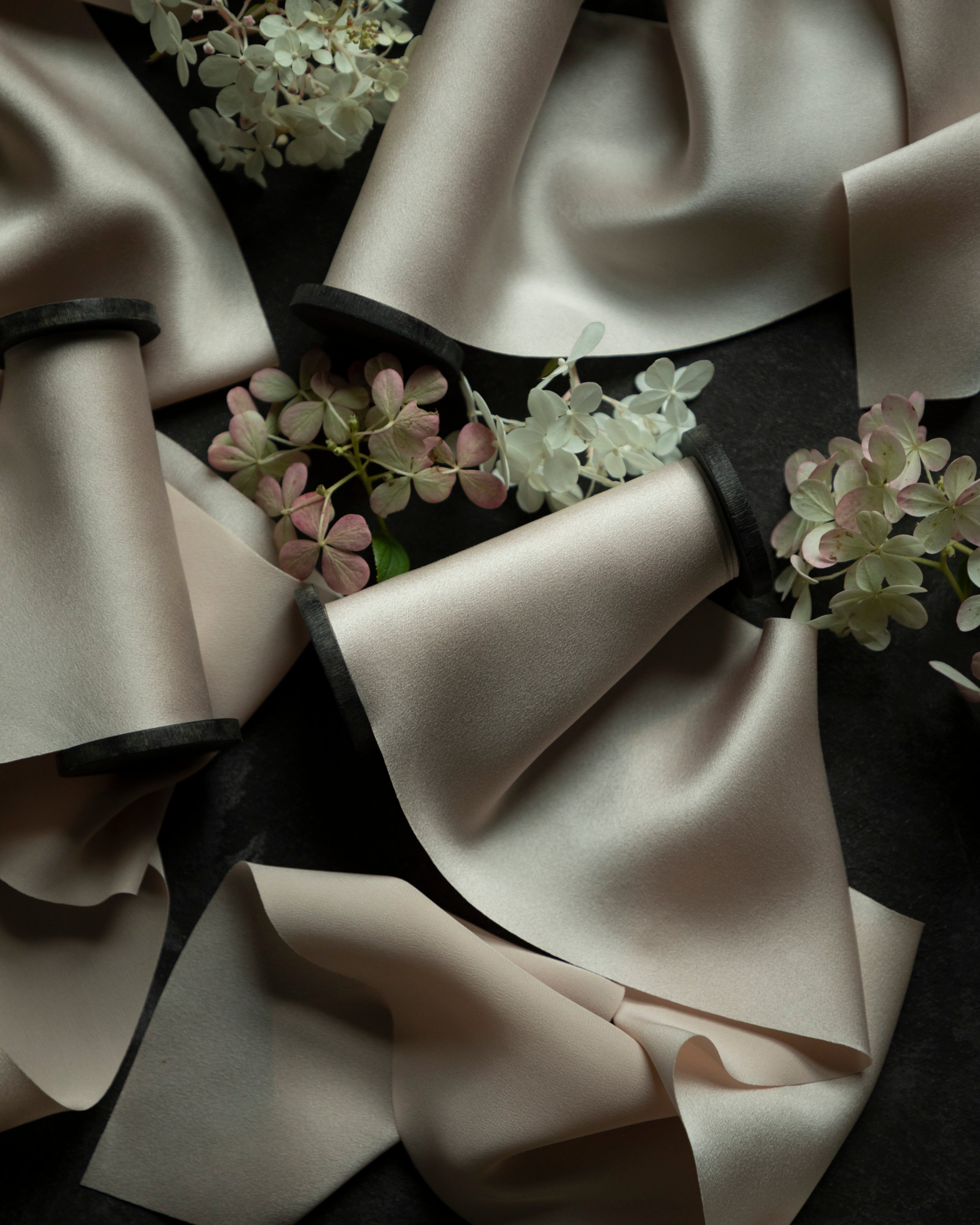 Blush silk satin ribbon spools with black bands, arranged on a dark background with decorative white and pink hydrangea flowers.