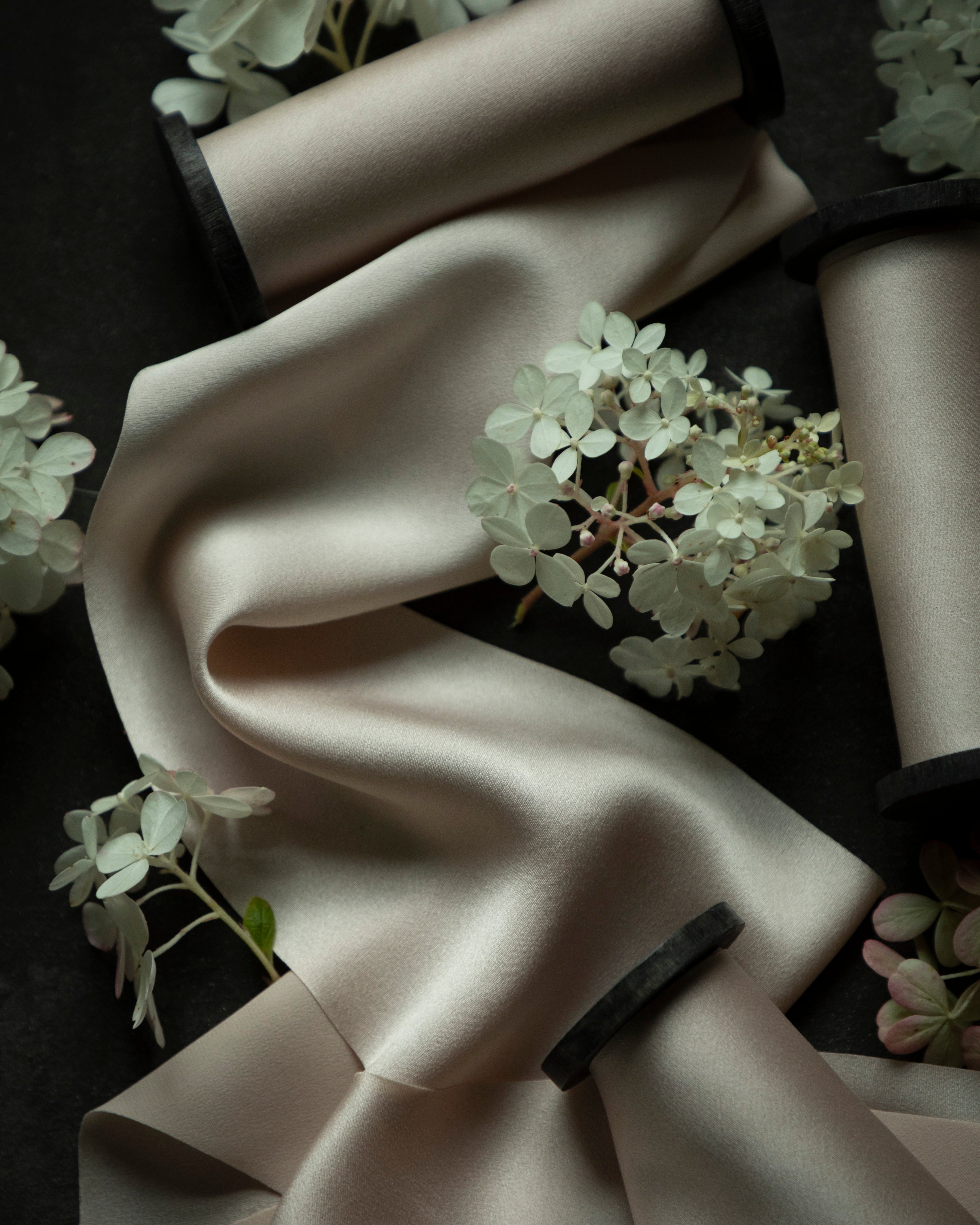 A draped satin fabric in a warm blush color, styled with white hydrangea flowers and wrapped around black spools on a dark background.