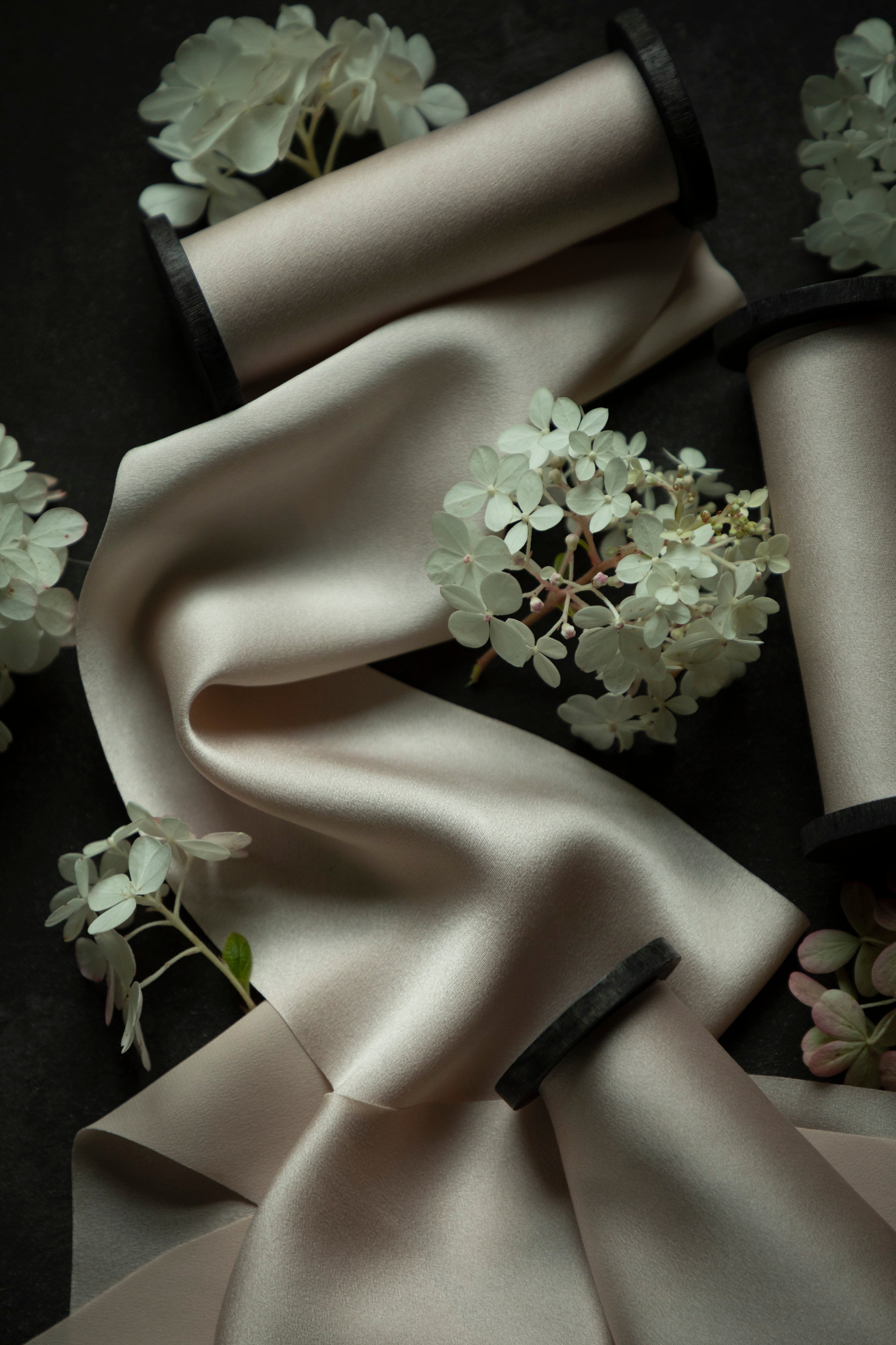 A draped satin fabric in a warm blush color, styled with white hydrangea flowers and wrapped around black spools on a dark background.