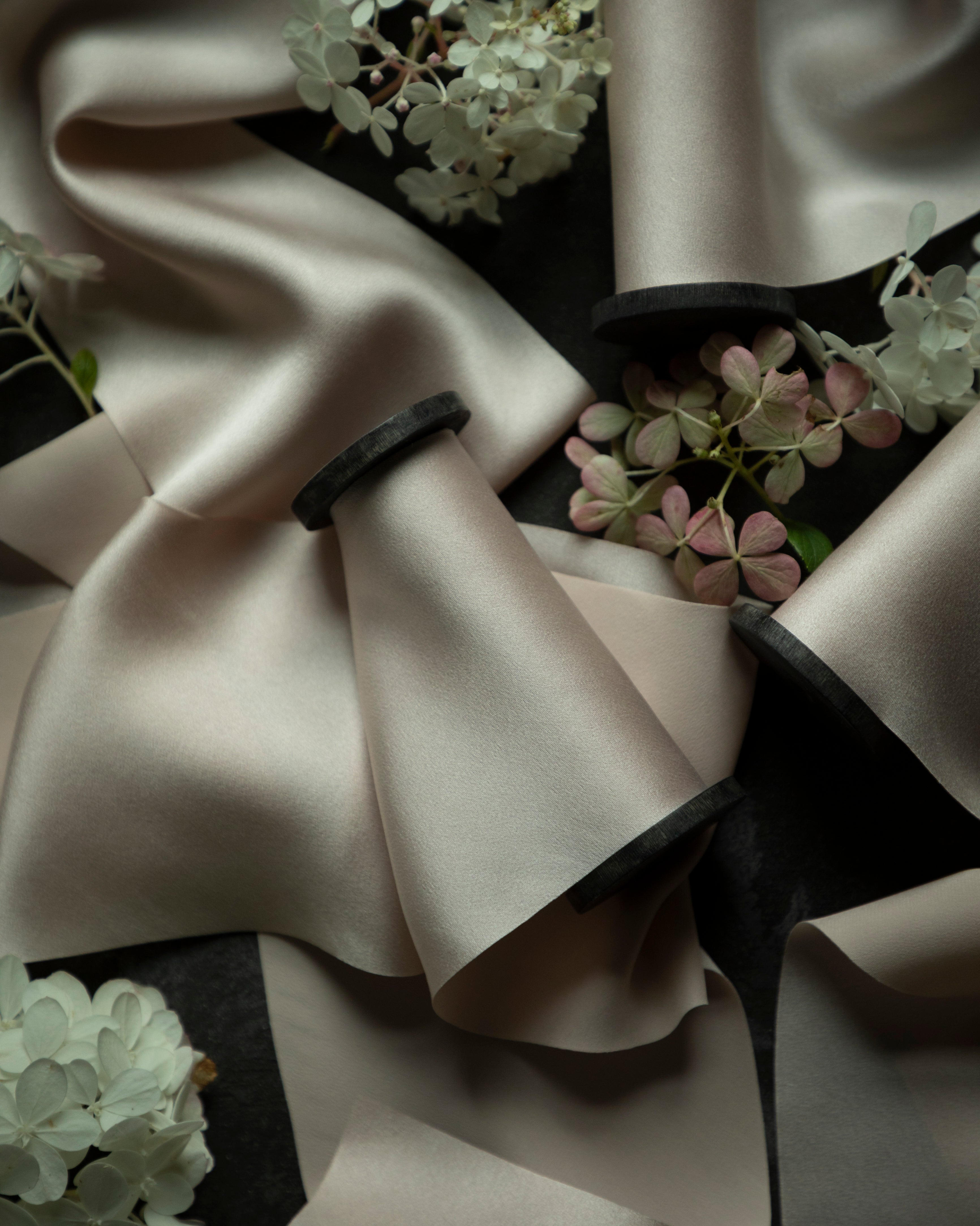 A close-up of draped bridal silk satin ribbons in a warm blush color, arranged with soft folds over a dark background and accented with small white and pink hydrangea flowers.