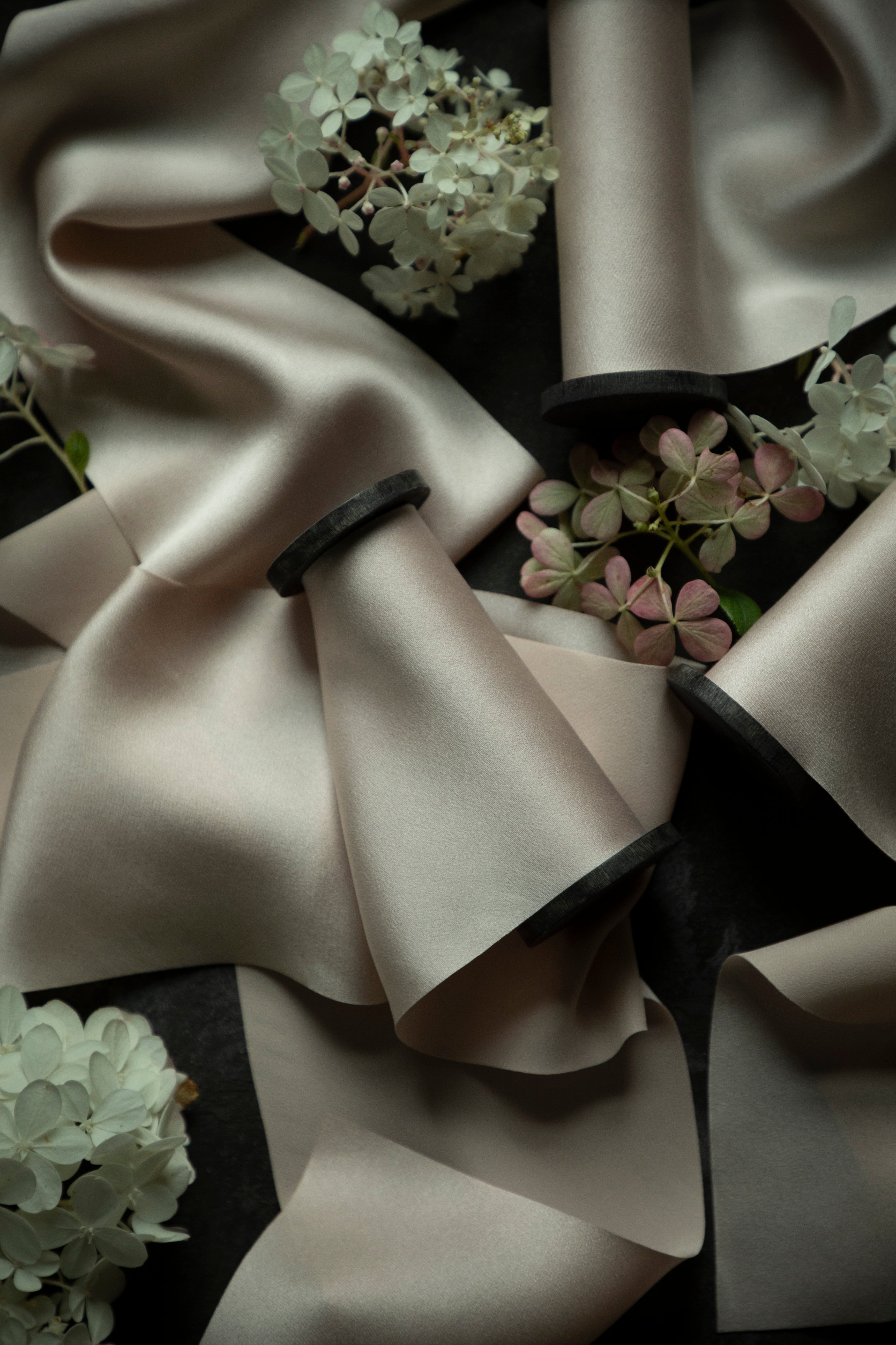 A close-up of draped bridal silk satin ribbons in a warm blush color, arranged with soft folds over a dark background and accented with small white and pink hydrangea flowers.
