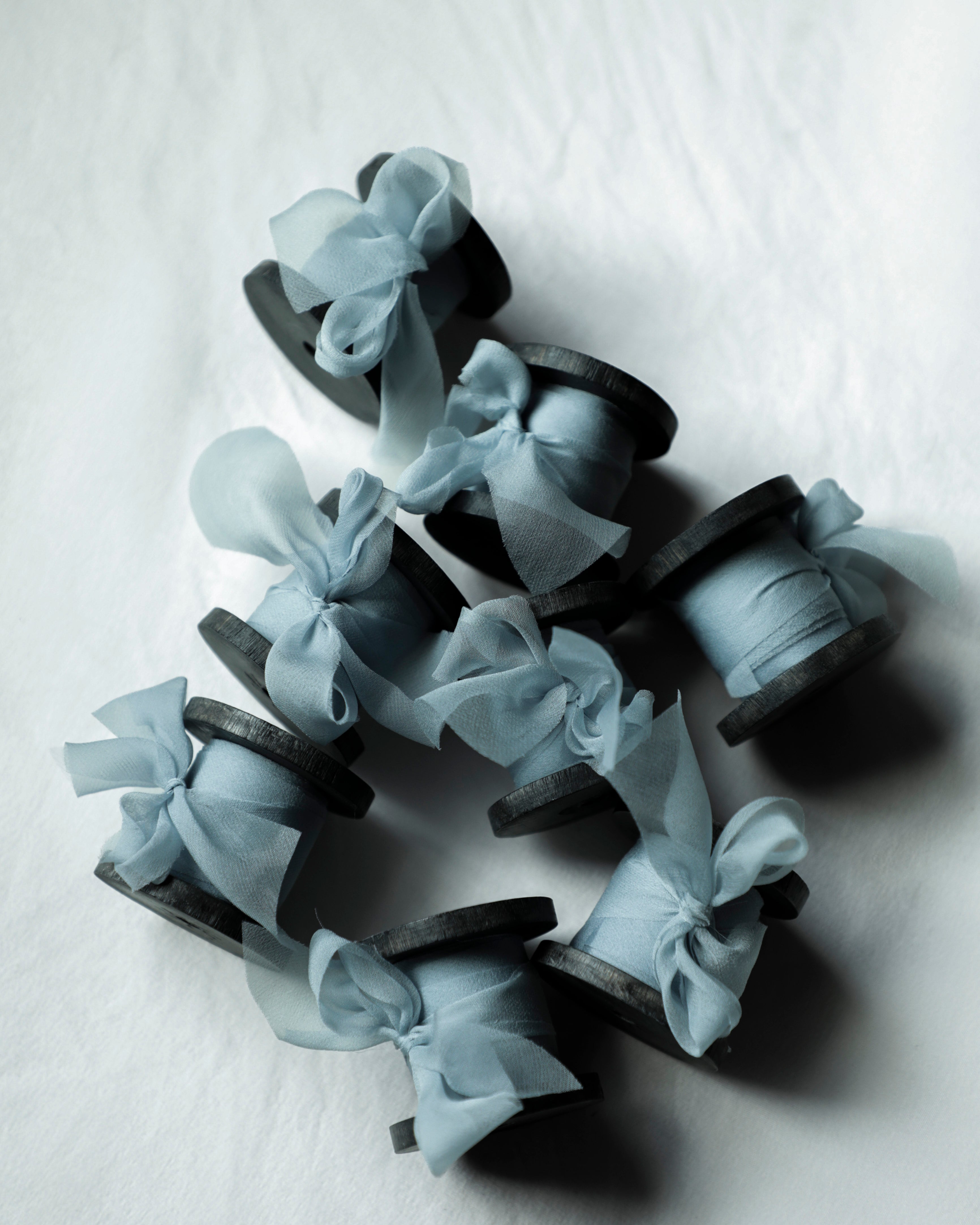 A group of small black wood spools wrapped with sheer pale blue ribbon bows, arranged on a light gray textured background.