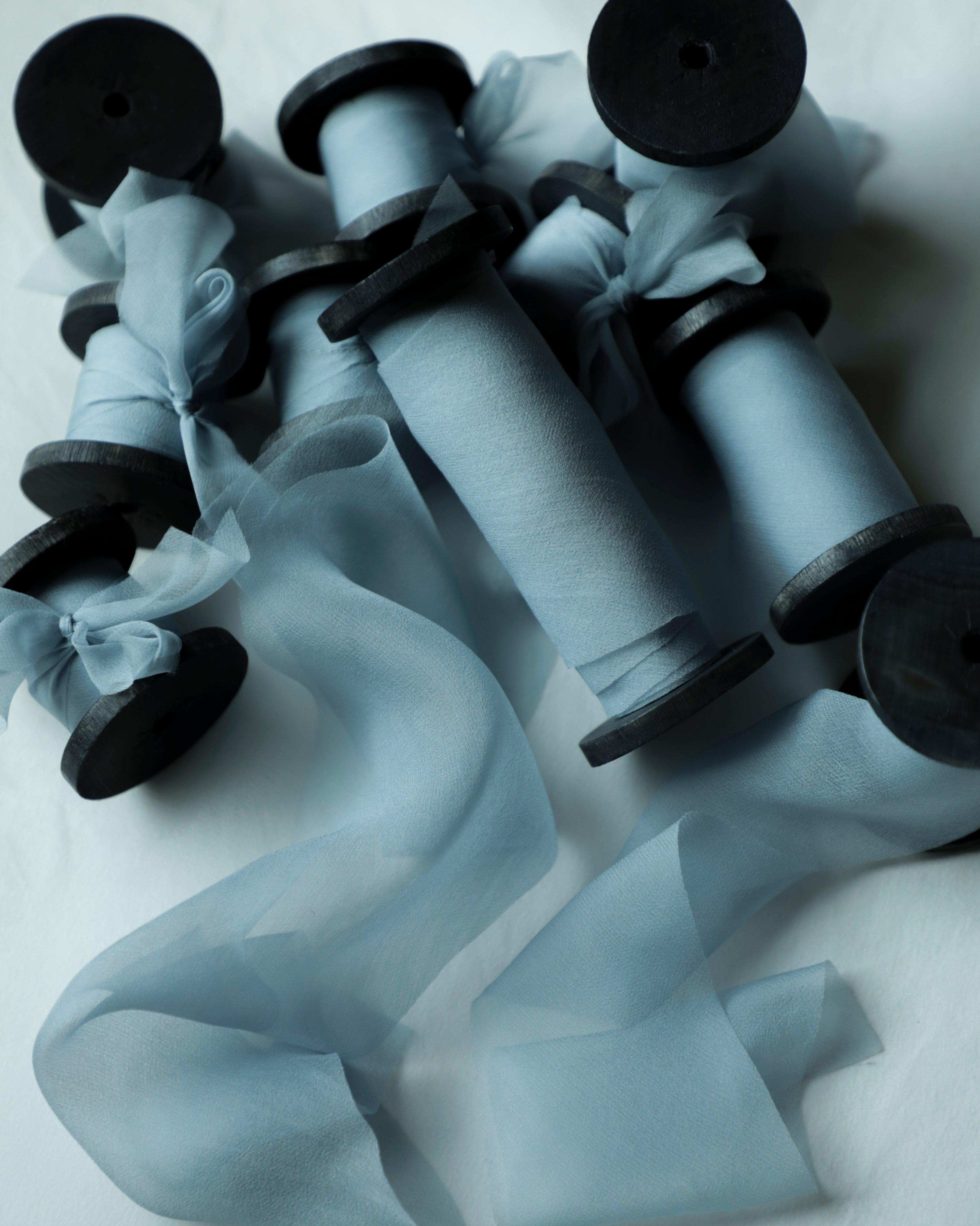 A set of light blue sheer organza ribbon spools on black wood spools, arranged on a white fabric background.
