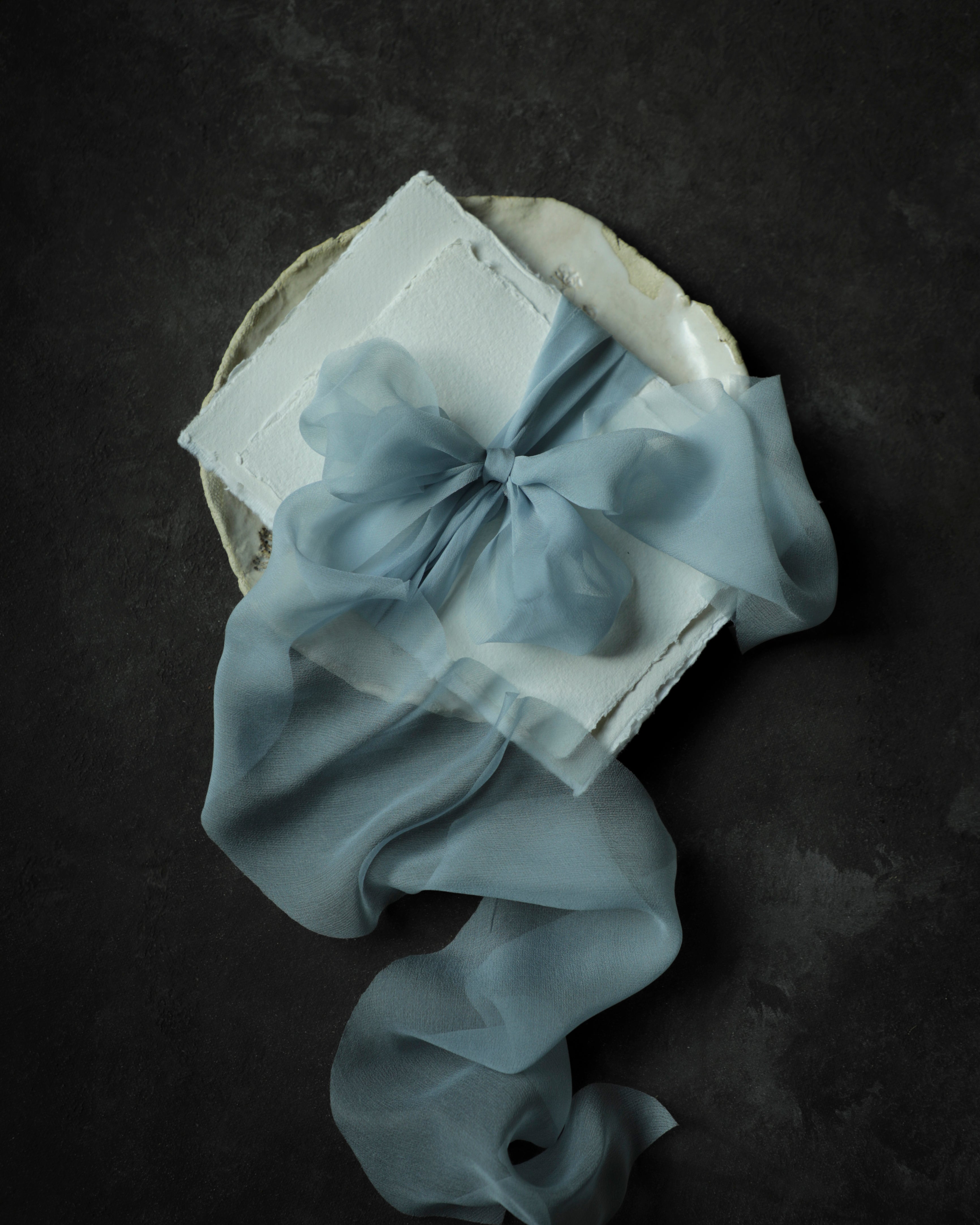 A light blue sheer organza ribbon tied into a large bow and draped over a white textured handmade deckle-edge paper envelope, presented on a dark textured background.