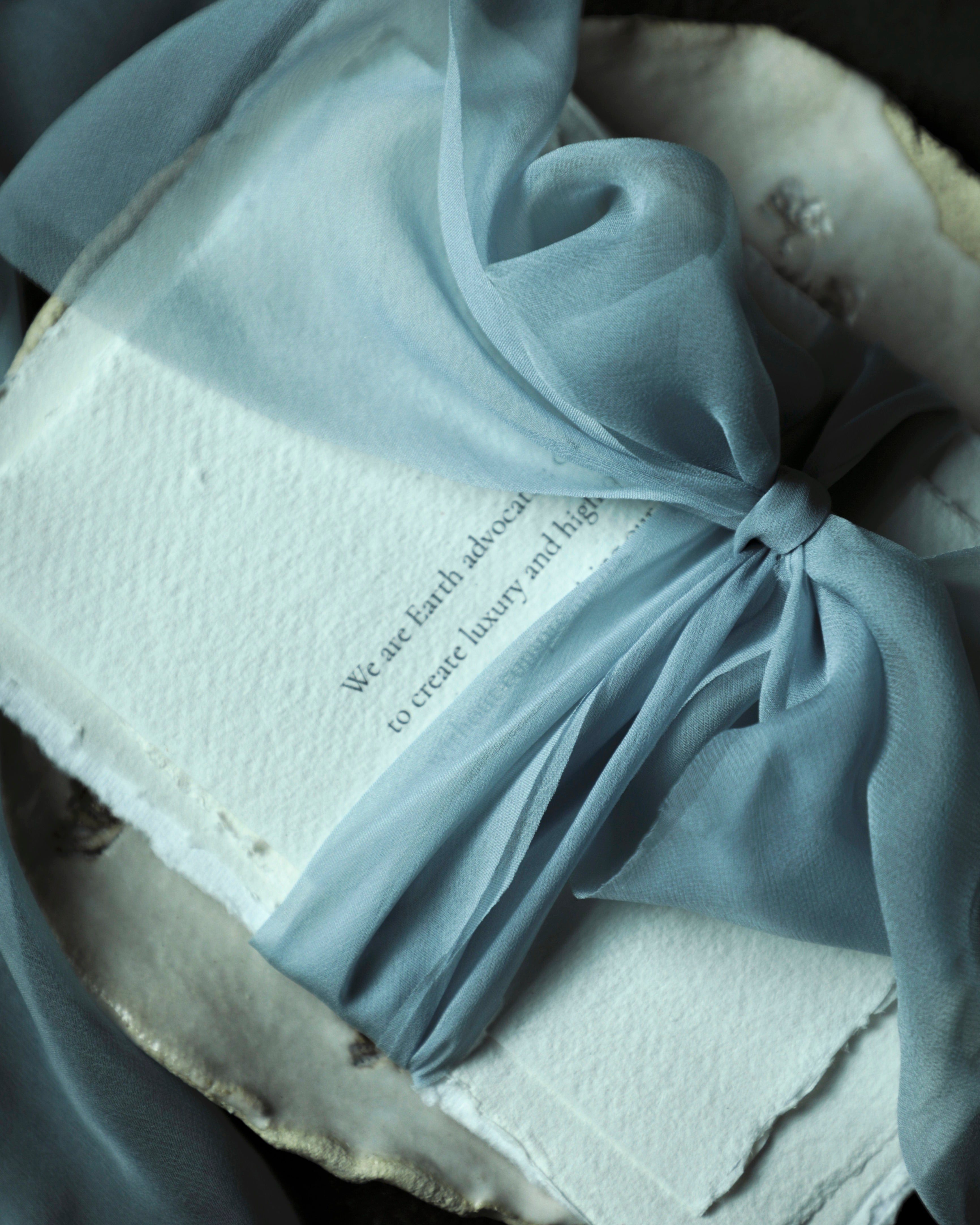 handmade deckled-edge paper wrapped with a frech blue sheer silk ribbon and tied with a big bow onto a handmade ceramic plate against a black backgraound.