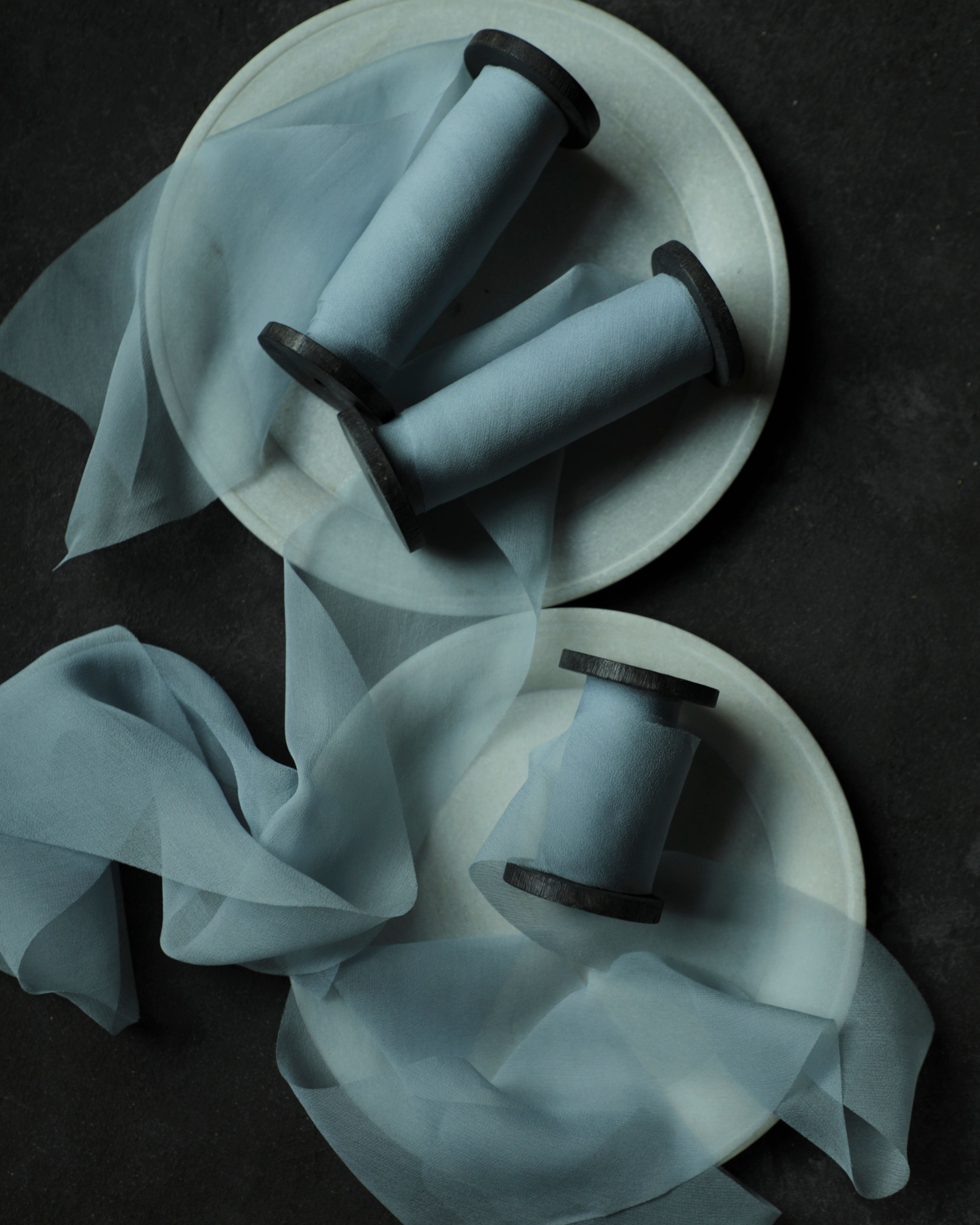 Three light blue spools of french blue silk ribbon arranged on white ceramic plates with sheer blue ribbons draped across a dark textured background.