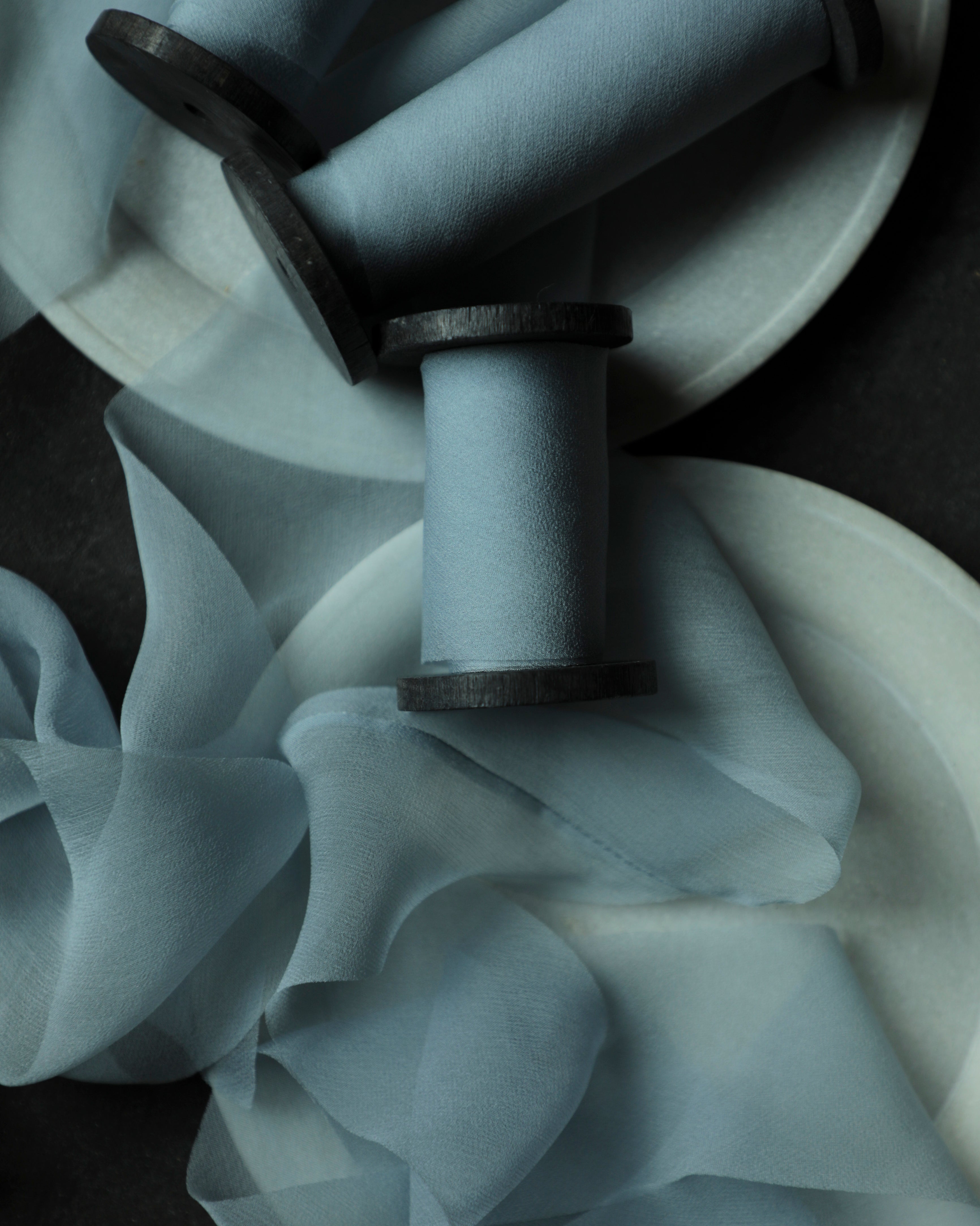 Light blue sheer silk ribbons draped over white circular plates, photographed in moody low light on a dark surface.