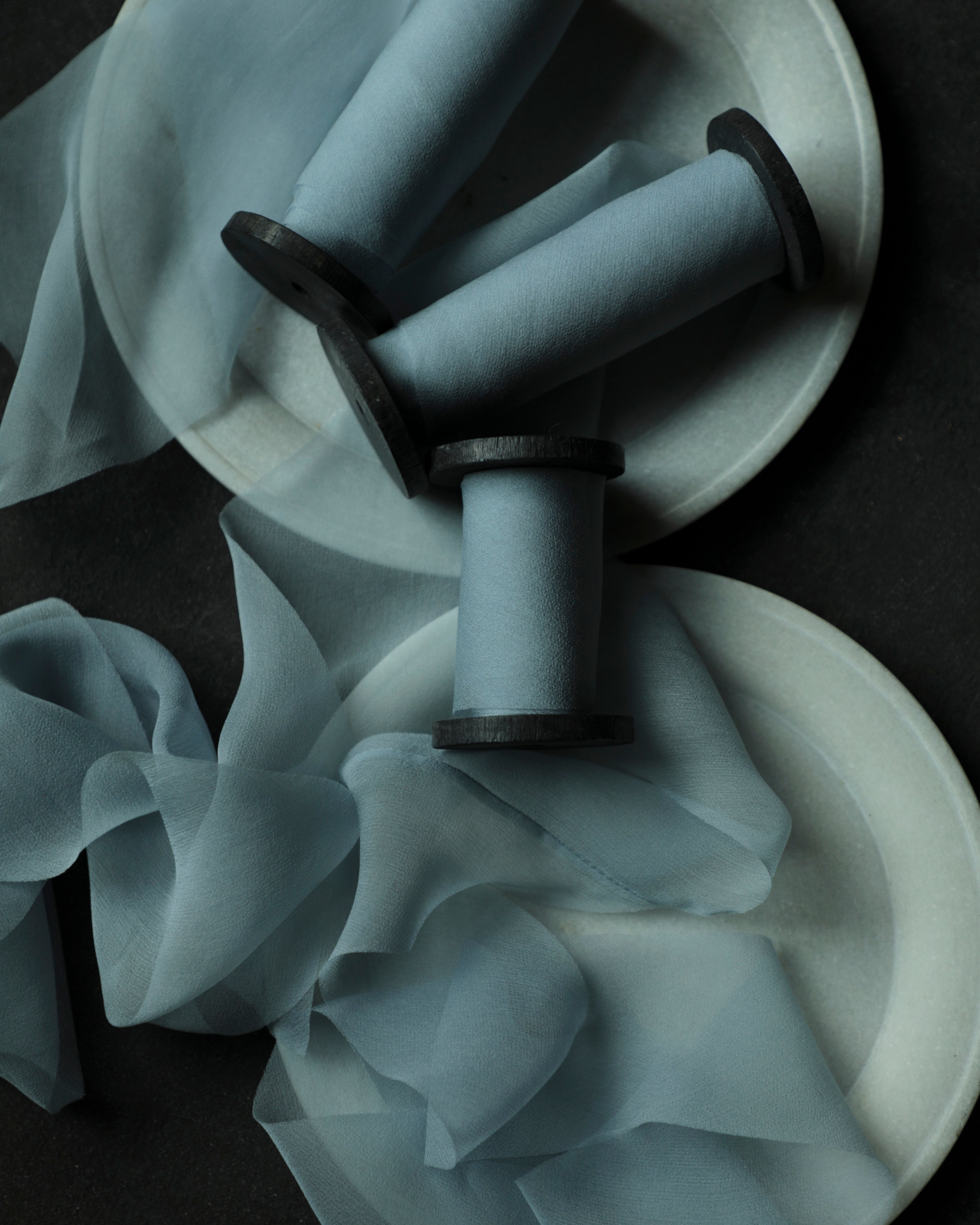 Three light blue organza ribbon spools with black wood spools, arranged on white plates with draped blue ribbon against a dark background.