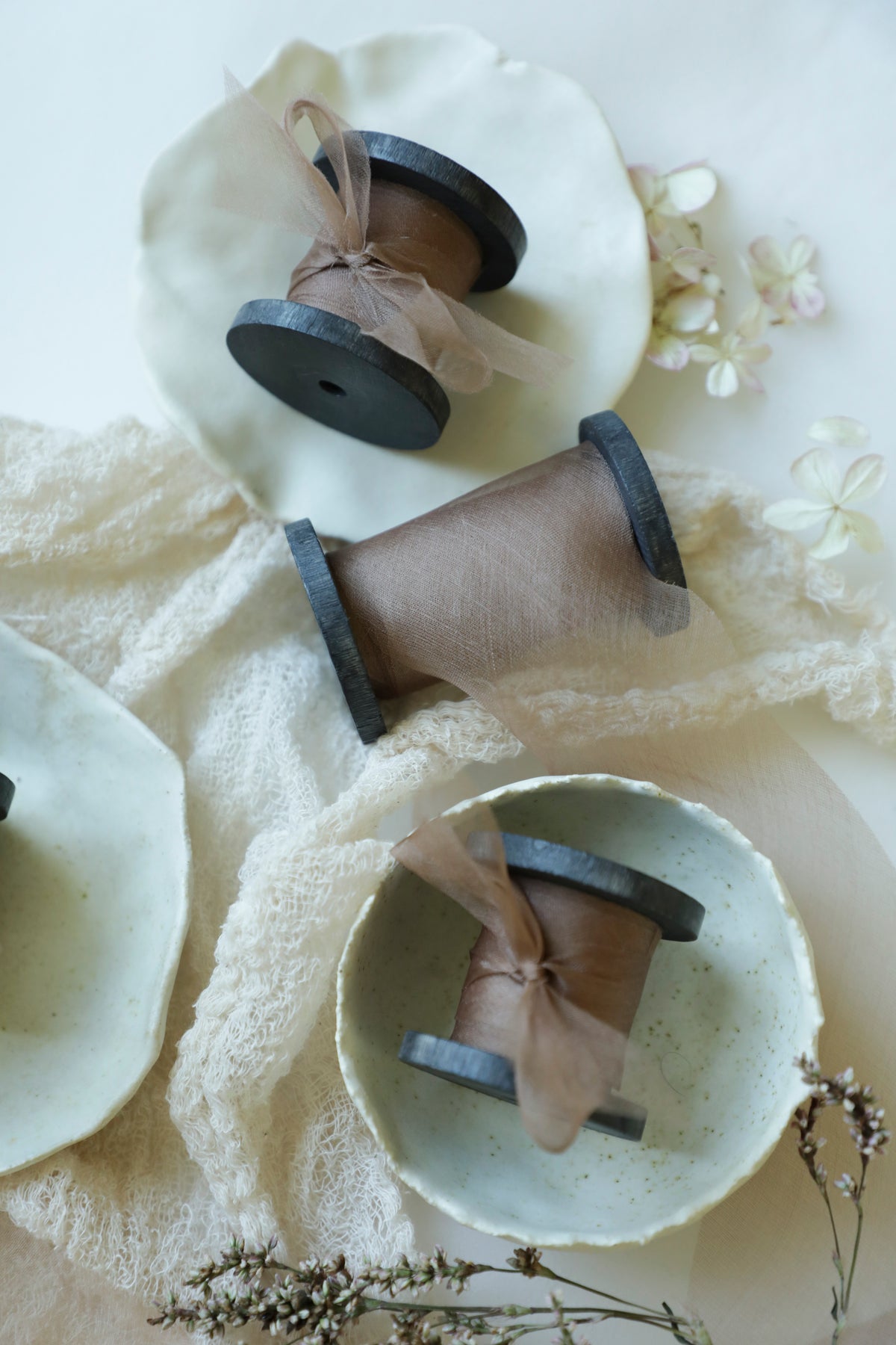 Three rustic wooden spools of silk ribbon wound on a black wood spool, styled on ceramic plates and a lace cloth.