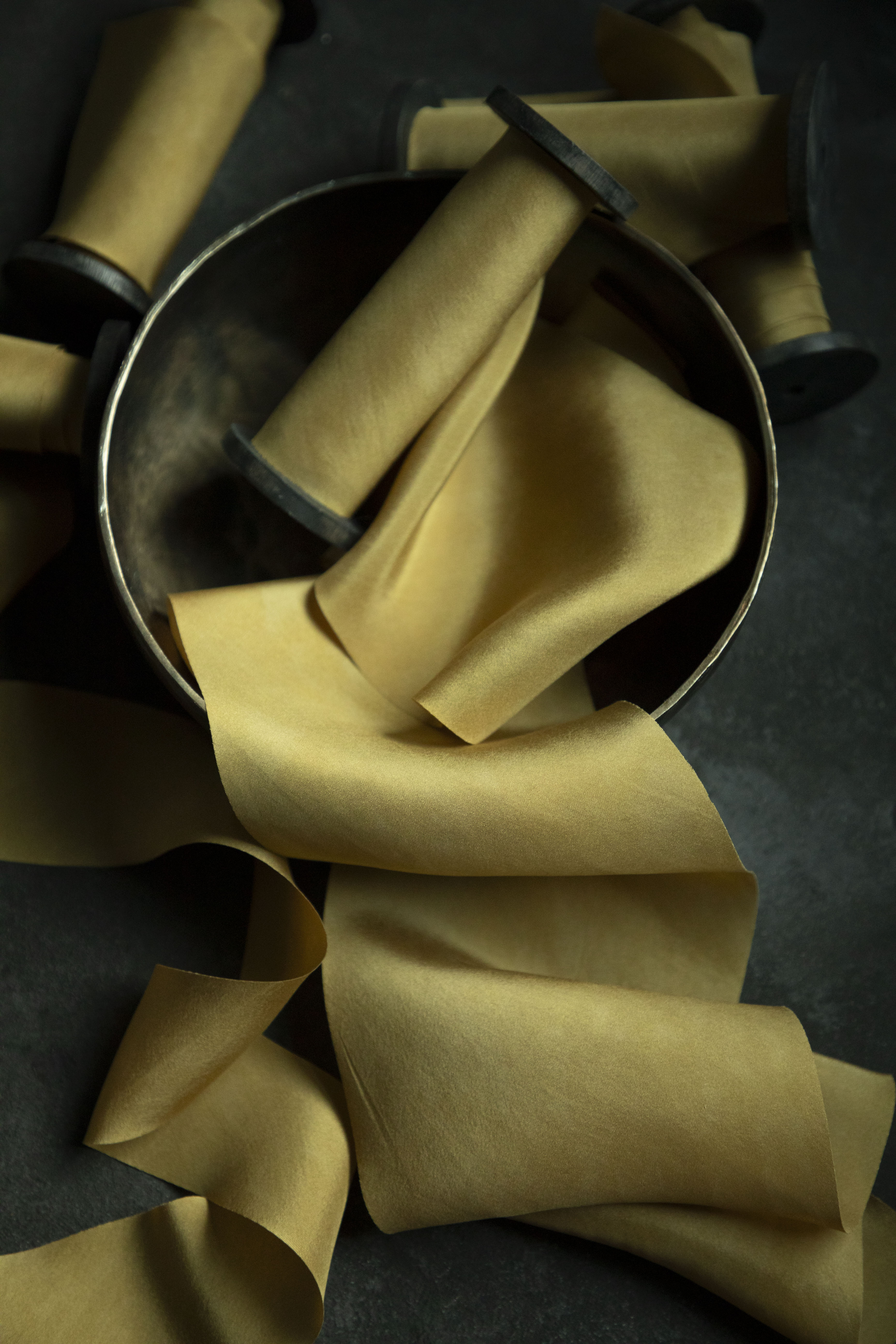 Yellow silk ribbons on a wooden spool on a dark surface with a metal bowl.