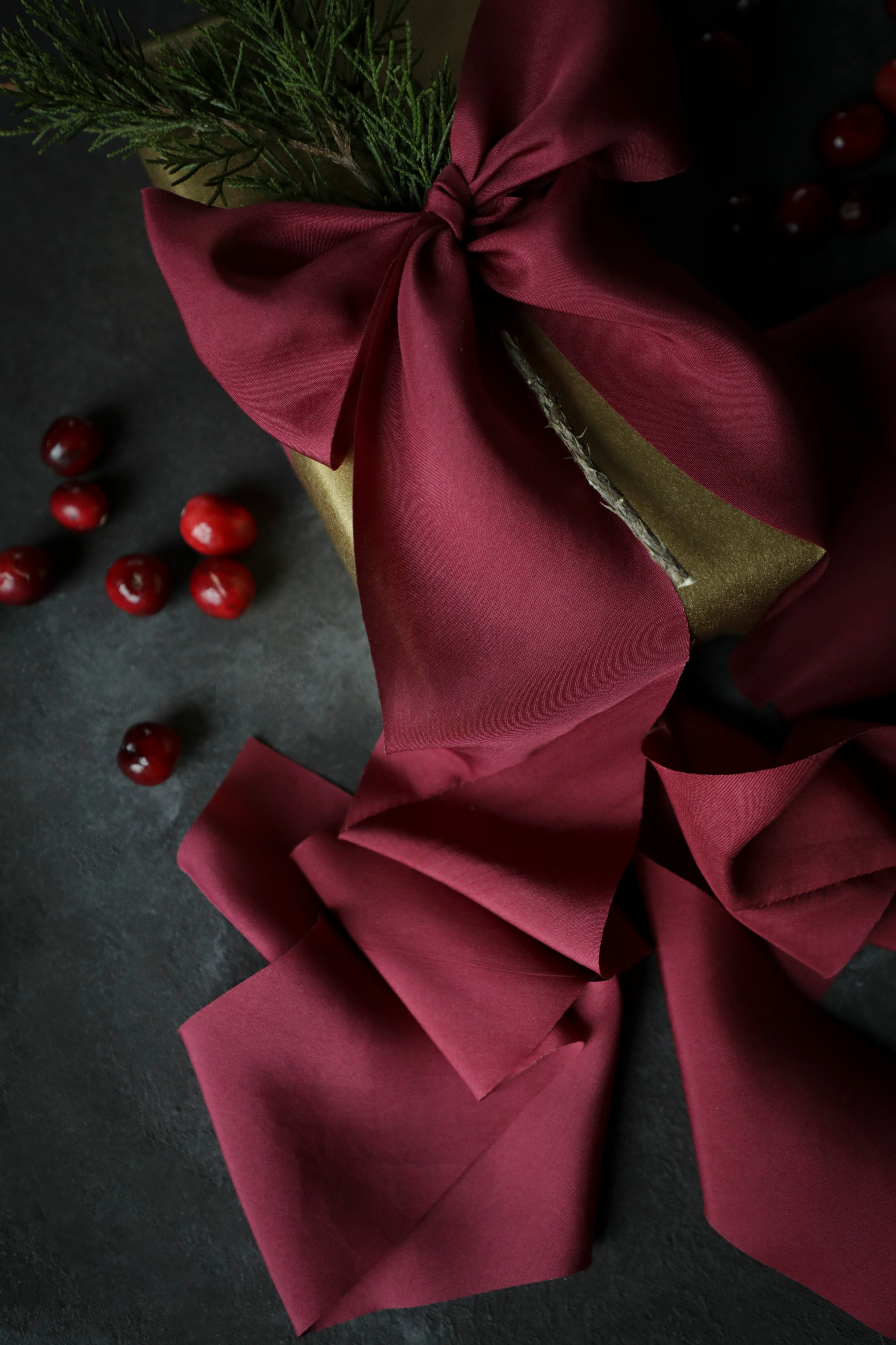 A piece of red silk ribbon with a bow, partially unrolled to show the smooth texture, placed on a dark background with clementines and greenery in the background.