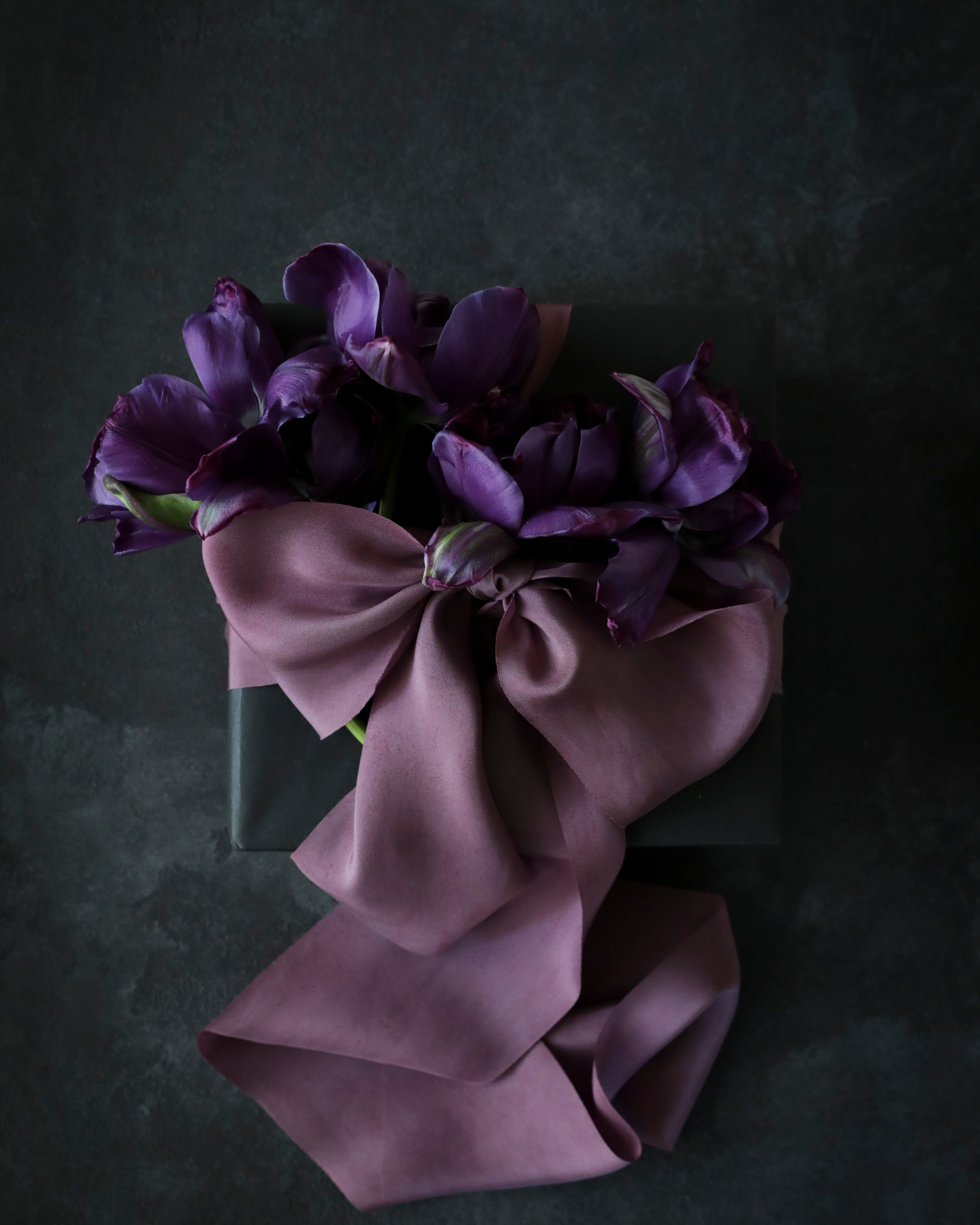 A deep mauve silk ribbon tied in a bow with a marbled pattern and speckles of plant matter, displayed on a dark background.