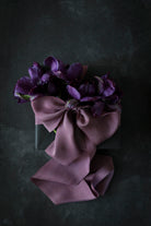 A deep mauve silk ribbon tied in a bow with a marbled pattern and speckles of plant matter, displayed on a dark background.