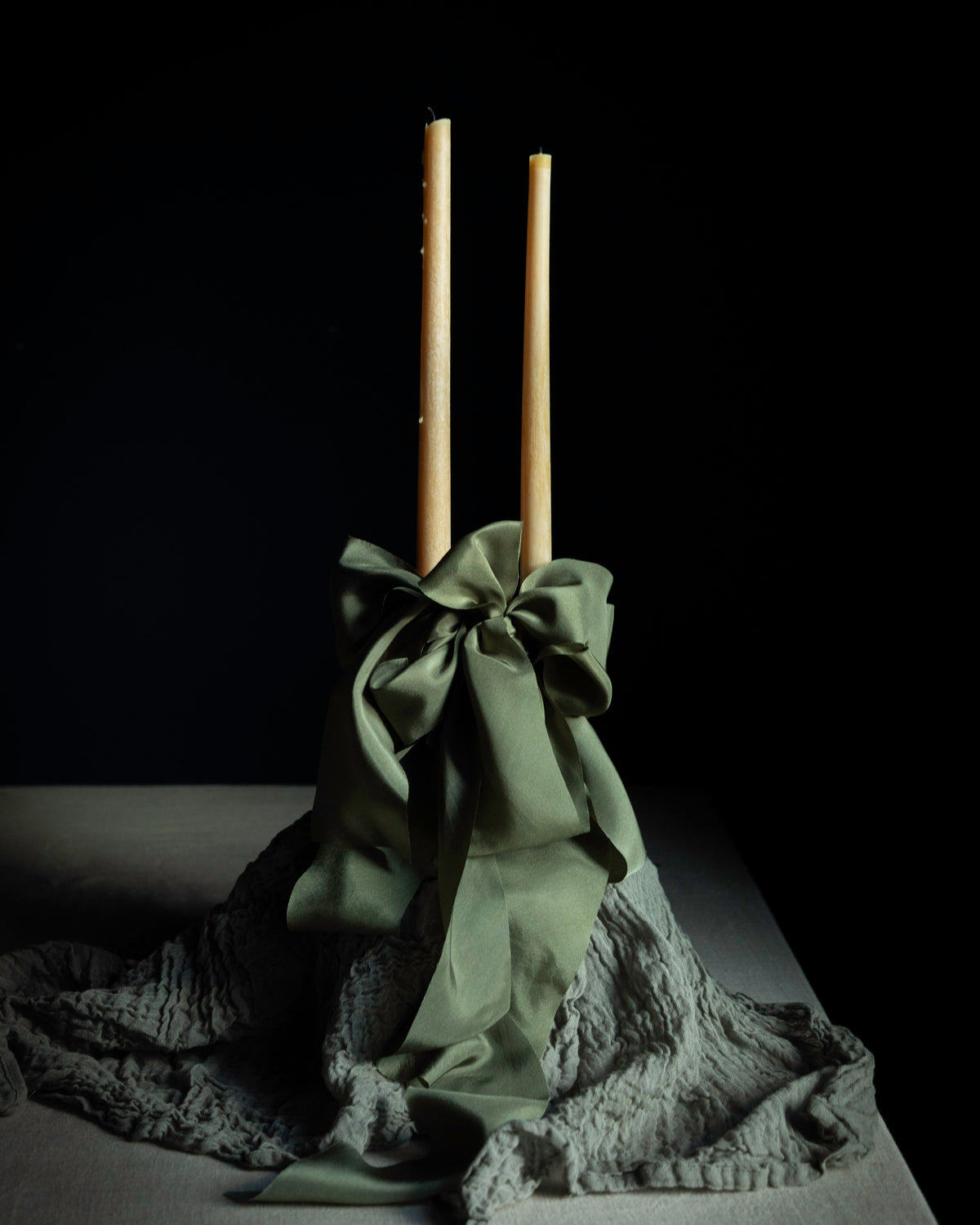 A decorative centerpiece featuring two tall natural wood taper candles standing upright in a gathered olive green fabric base with a large bow, draped over a textured gray fabric on a dark table against a black background.