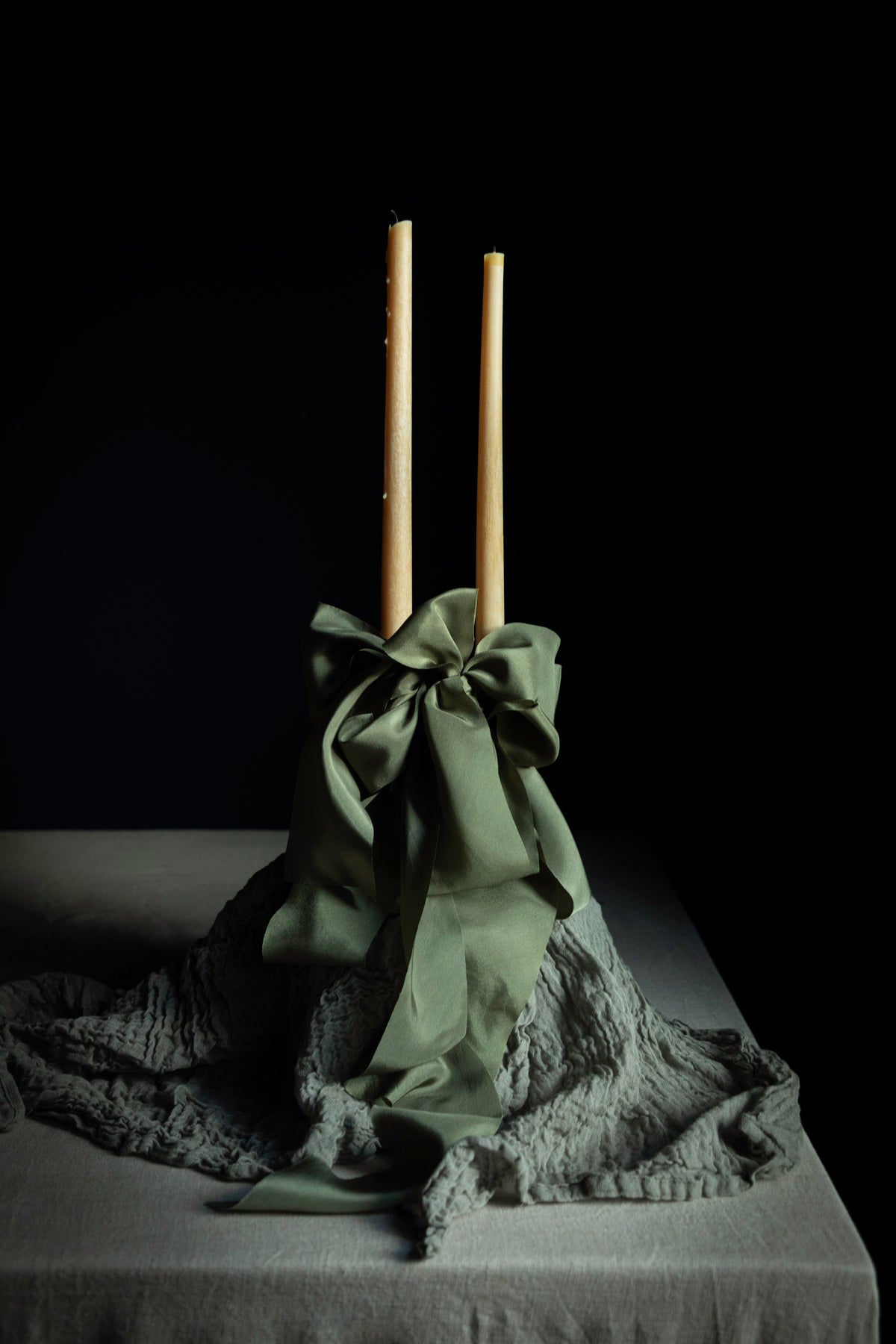 A decorative centerpiece featuring two tall natural wood taper candles standing upright in a gathered olive green fabric base with a large bow, draped over a textured gray fabric on a dark table against a black background.