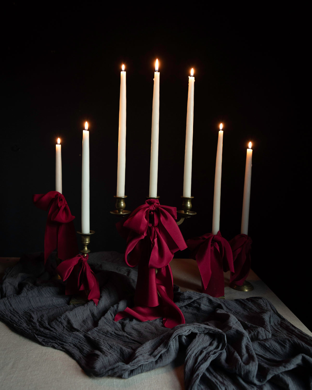 Candles display draped with red ribbon bows on layered gray table textiles against a dark background